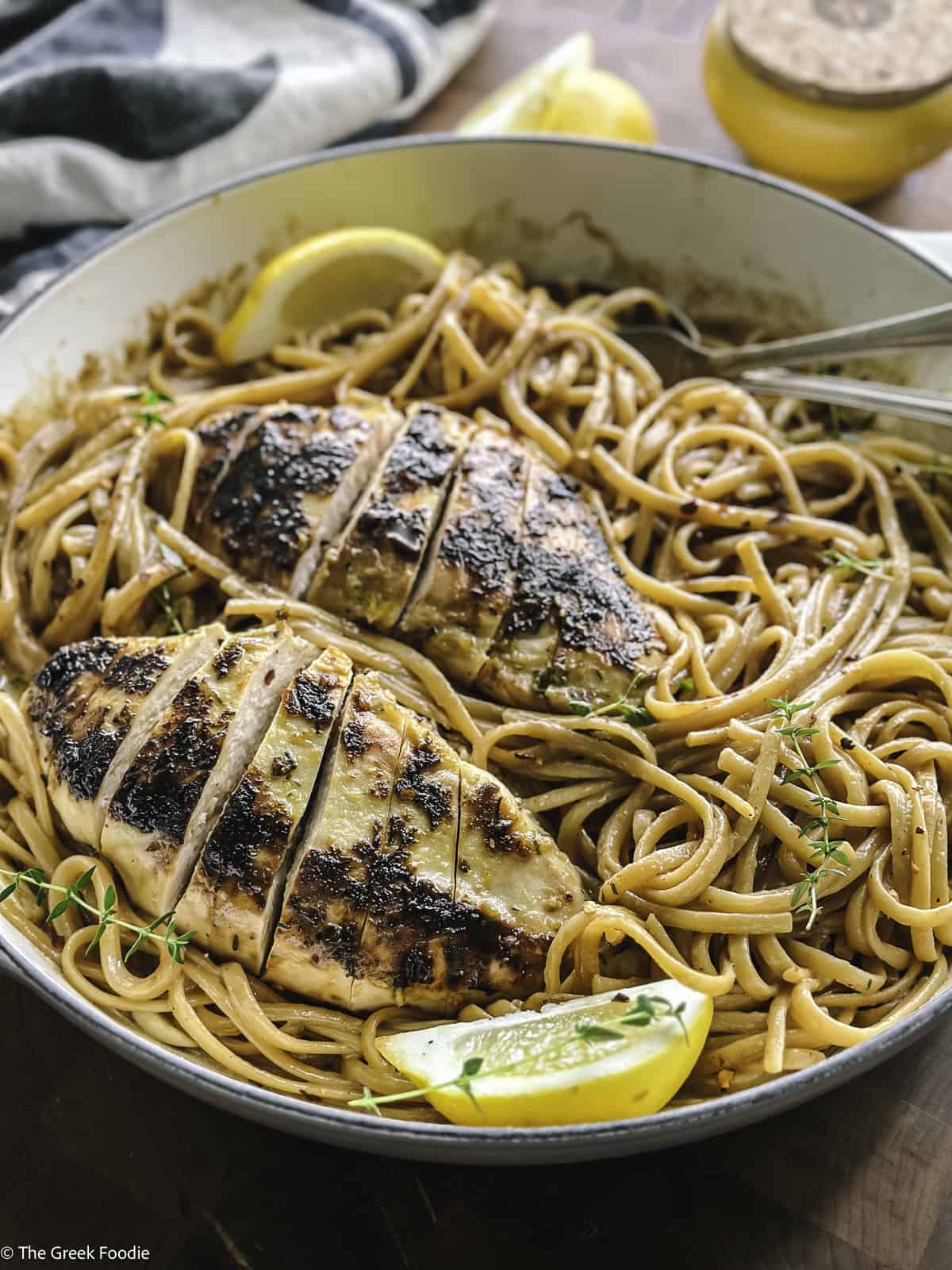 A plate with lemon chicken pasta, utensils and a lemon wedge.