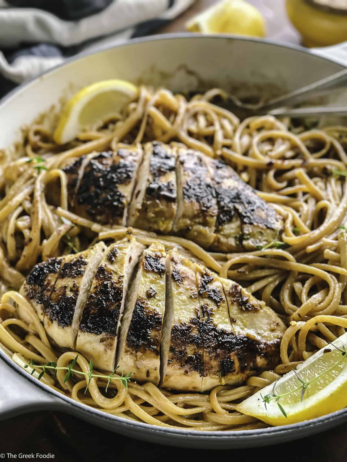 A plate with lemon chicken pasta, utensils and a lemon wedge.