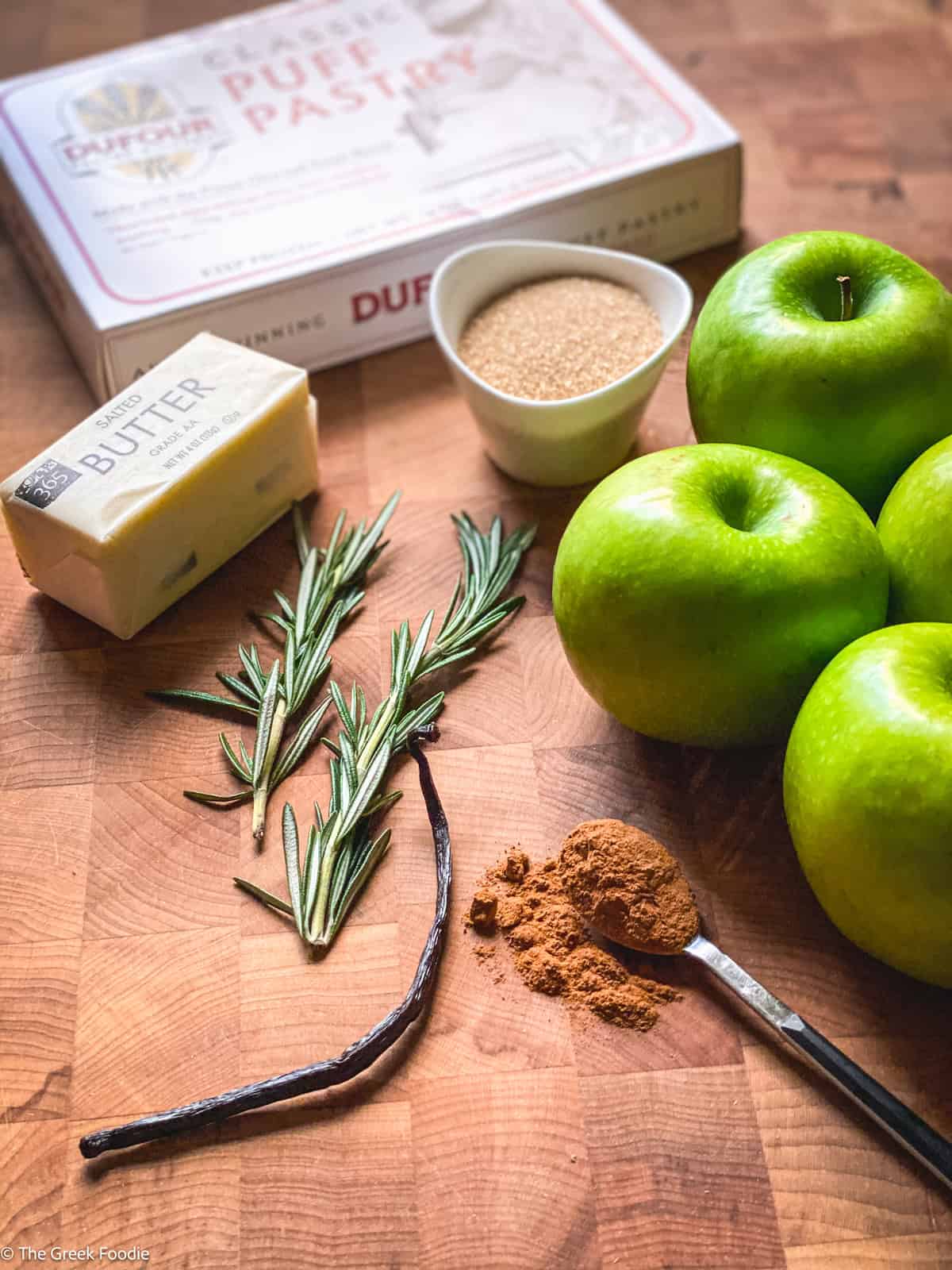 Puff pastry packet, apples, a vanilla bean, butter, a cup with sugar and some rosemary sprigs on a cutting board.