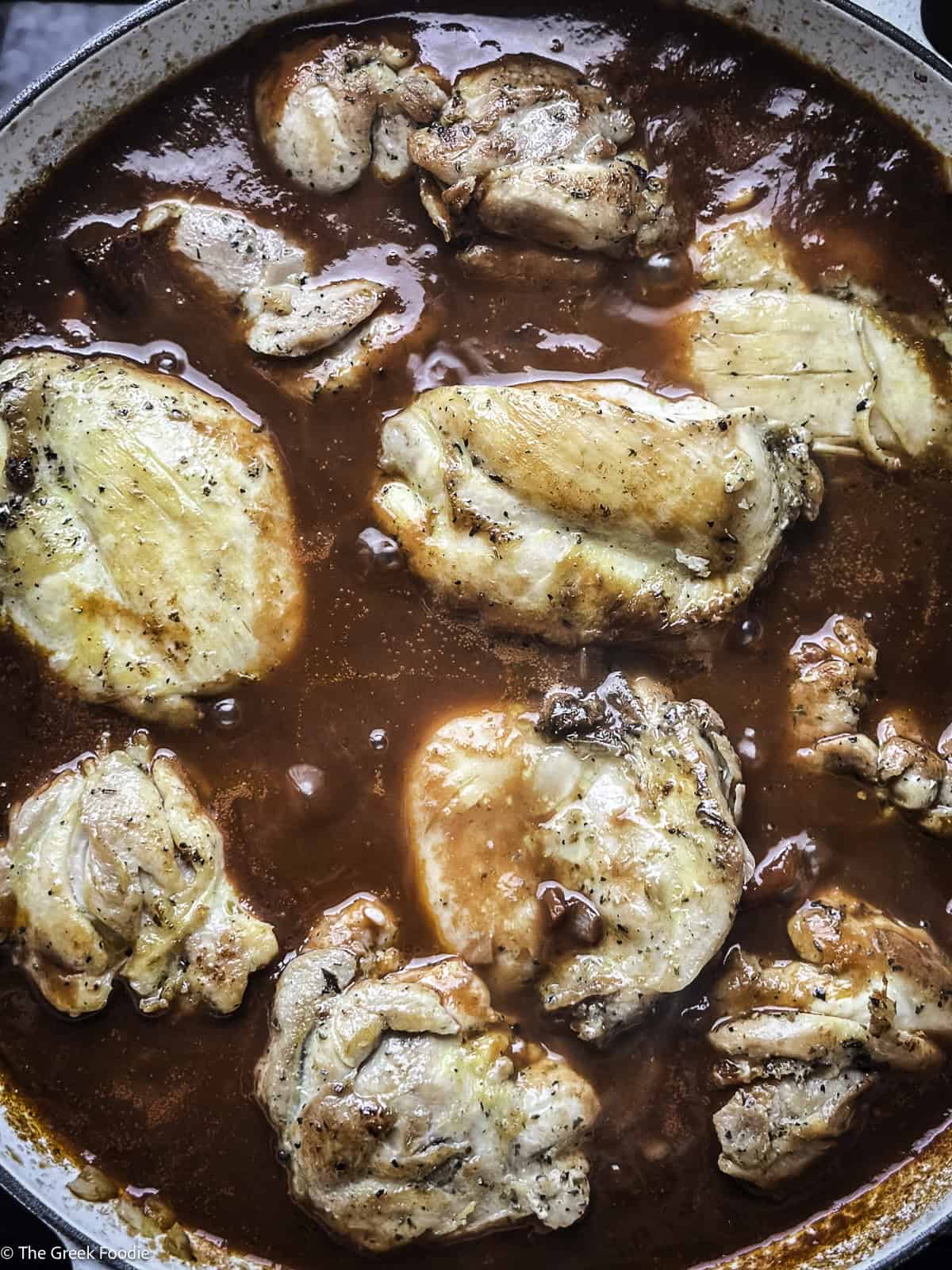 Wine sauce and chicken thighs cooking in a white skillet.