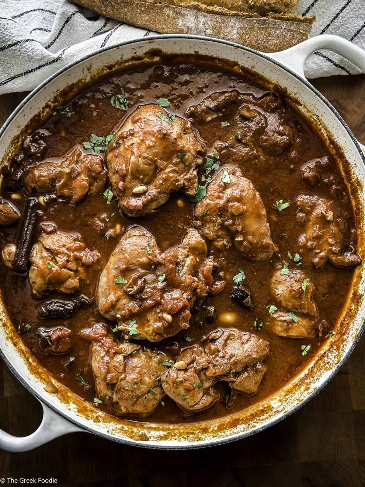 A white skillet with Greek chicken Kapamas stew on a table with a loaf of bread and a pepper mill.