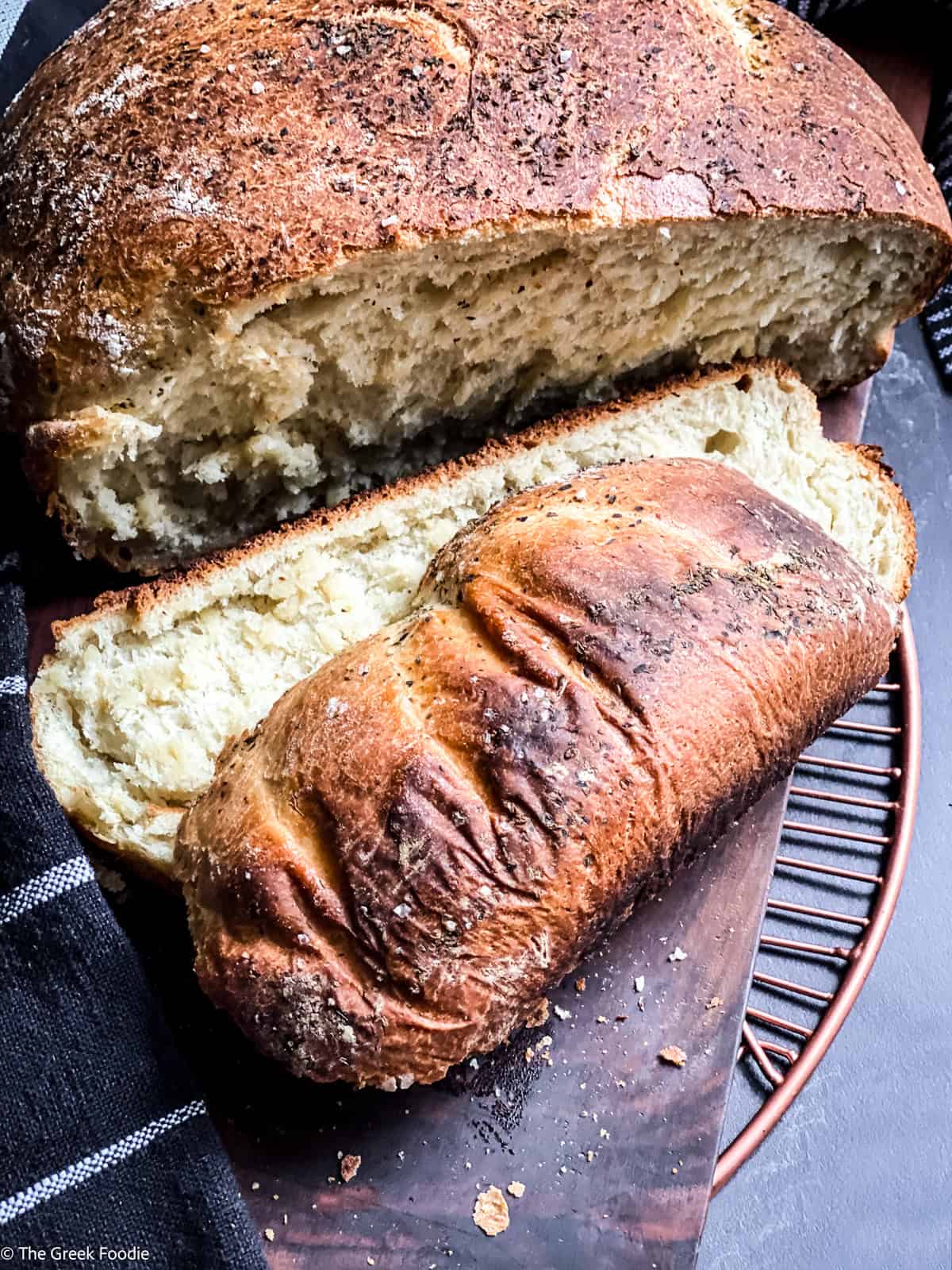 A loaf of Greek bread with two slices cut on a wooden cutting board. A metal coaster under it.