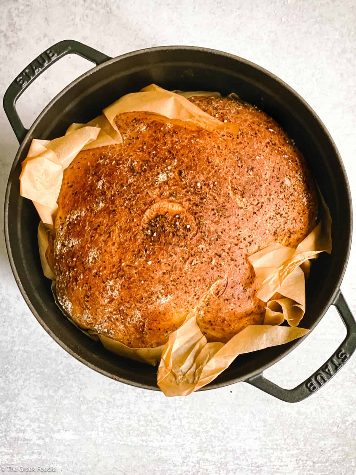 A black dutch oven with parchment paper and baked bread loaf in it on a cement surface.