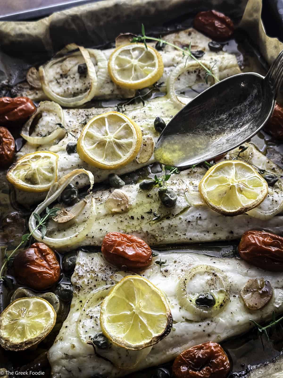 Roasted branzino fillets with cherry tomatoes and lemon slices in a baking tray, a spoon drizzling ladolemono on top.