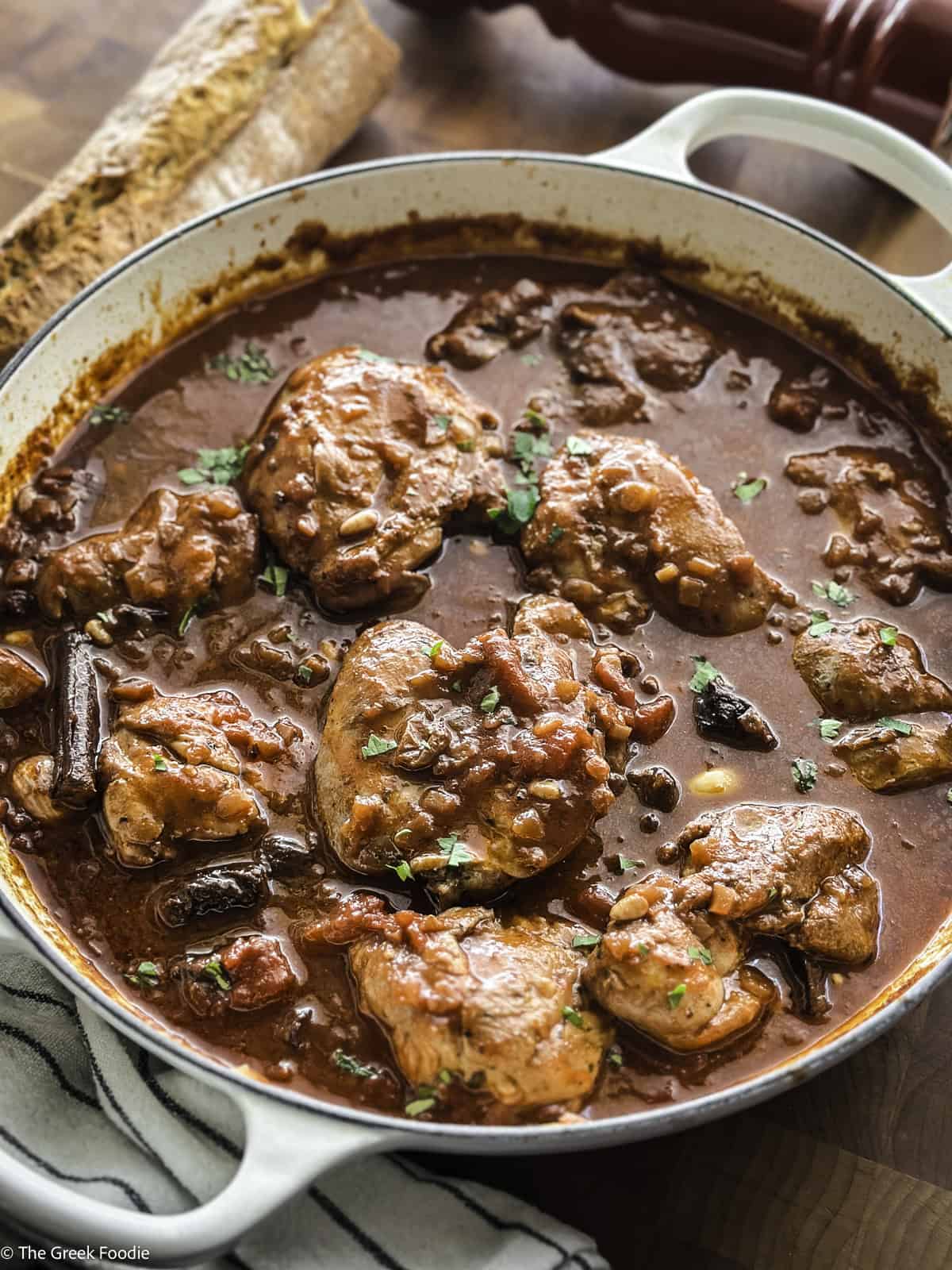 Chicken kapama in a white skillet on a table with a loaf of bread and a cloth napkin.