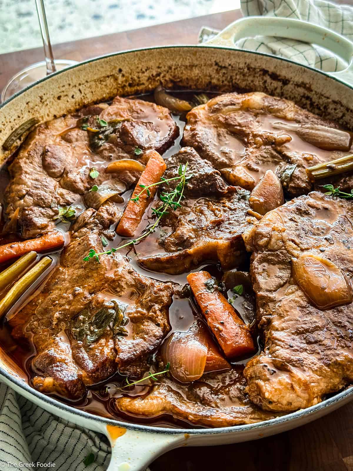 Pork shoulder slices in a dutch oven skillet with carrots and celery in a wine sauce, on a table.