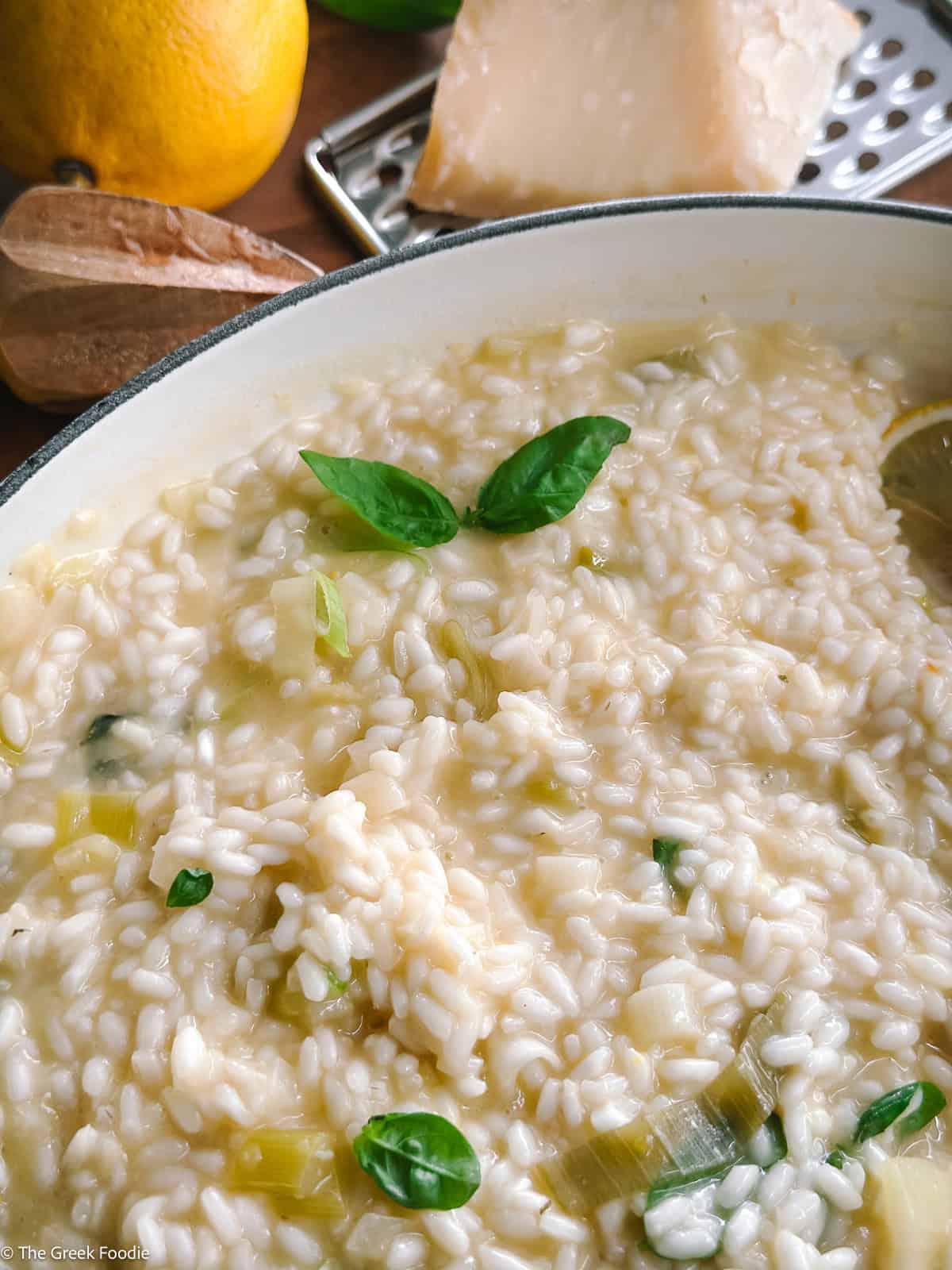 Creamy lemon herb risotto and a spoon in a white large saucepan, a lemon, parmesan at the back.