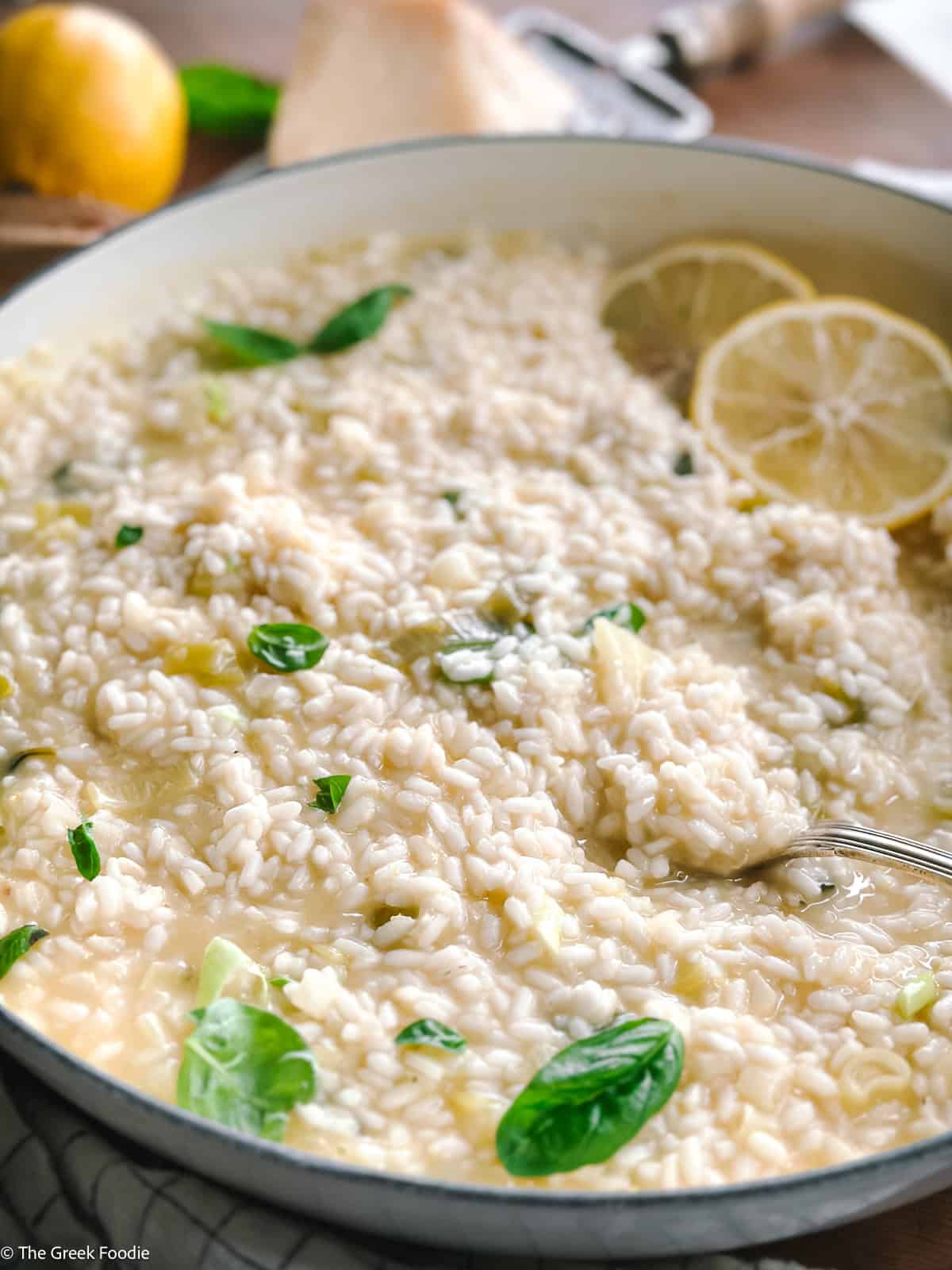 Creamy lemon herb risotto and a spoon in a white large saucepan, a lemon, parmesan at the back.