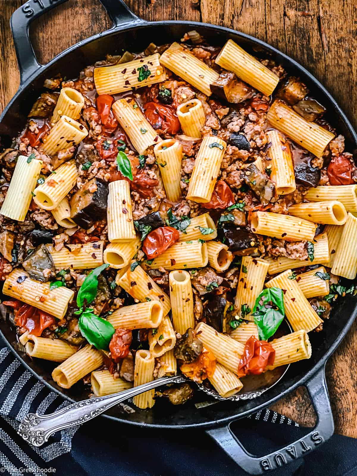 A cast iron skillet with eggplant pasta, utensils and a cloth napkin under it on a wooden table.