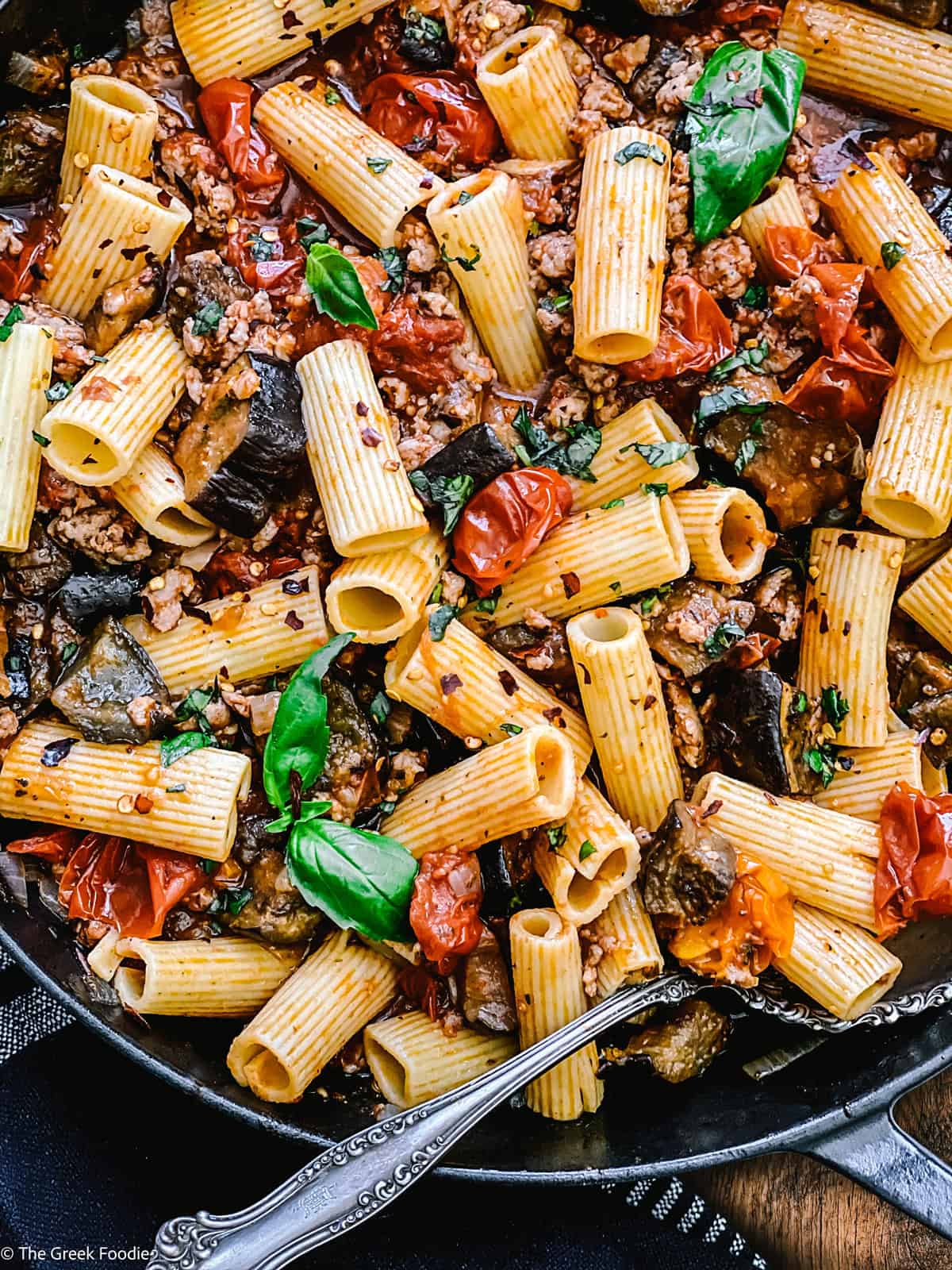 A cast iron skillet with eggplant pasta, utensils and a cloth napkin under it on a wooden table.