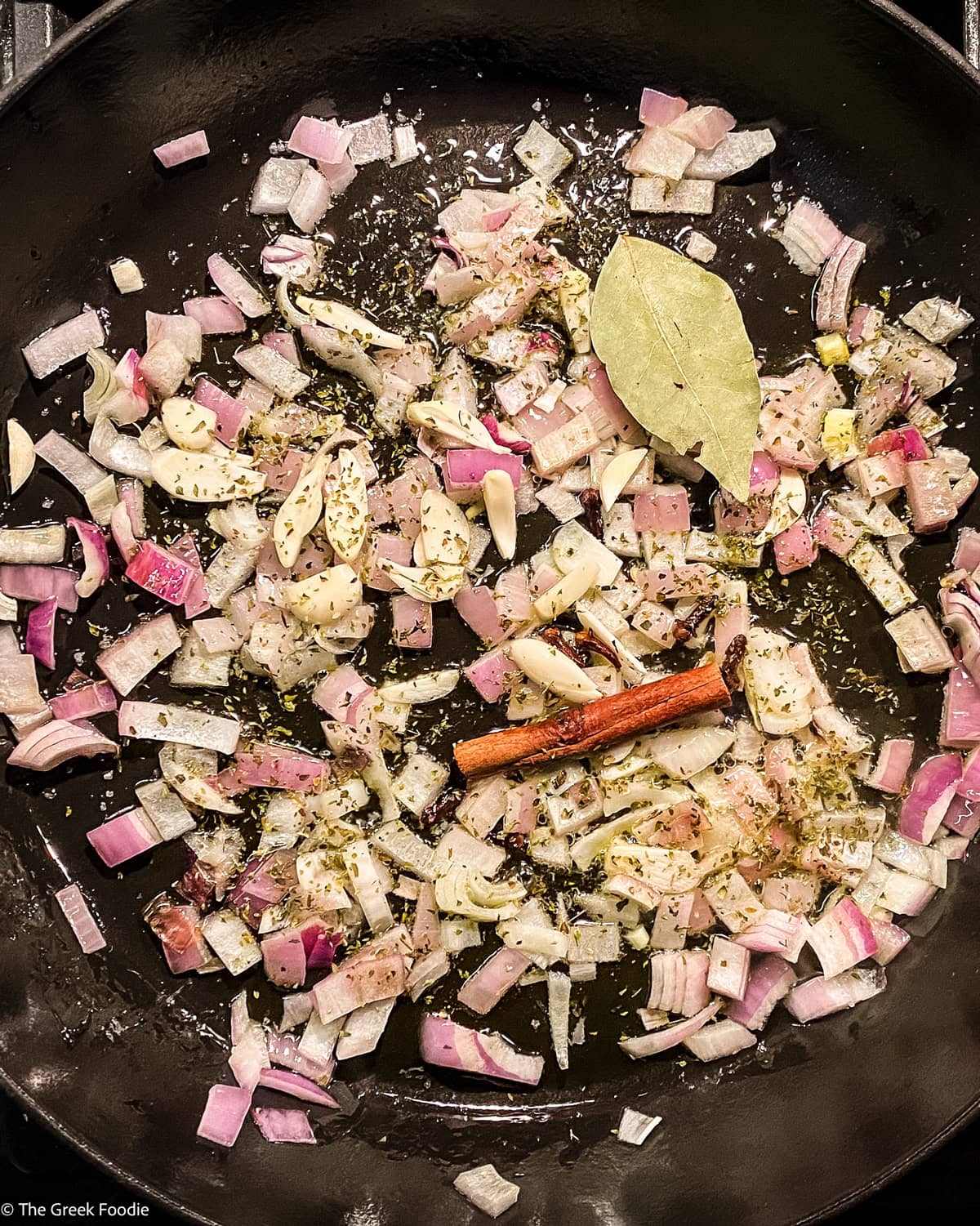 Onion, a bay leaf, a cinnamon stick and garlic cooking in a skillet.