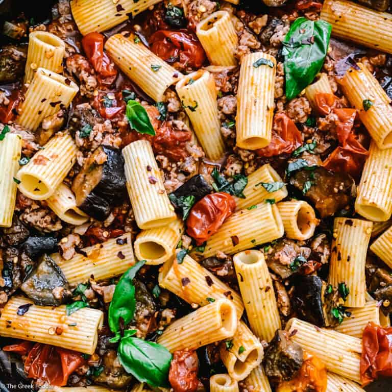 A cast iron skillet with eggplant pasta, utensils and a cloth napkin under it on a wooden table.