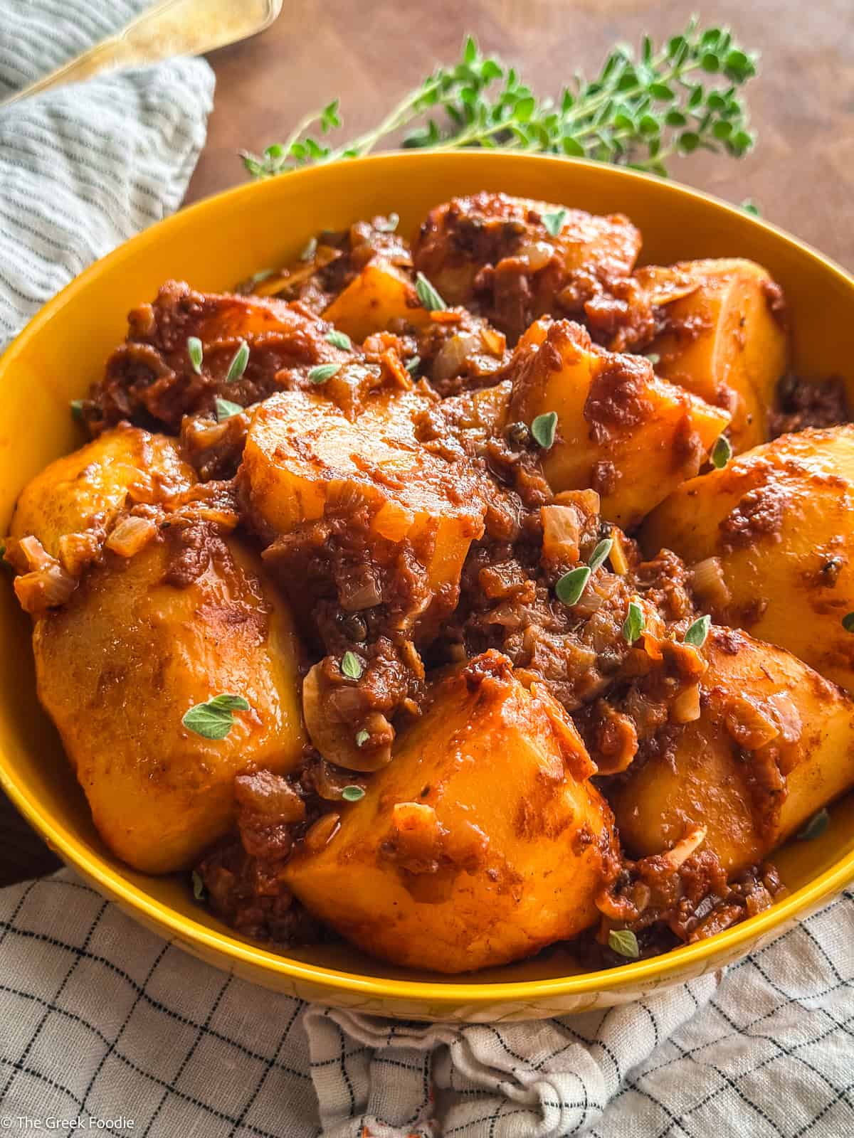 A yellow plate with large potato pieces in a red sauce, a Greek potato stew-patatew yahoo, with utensils, some herbs on the table and a cloth napkin.