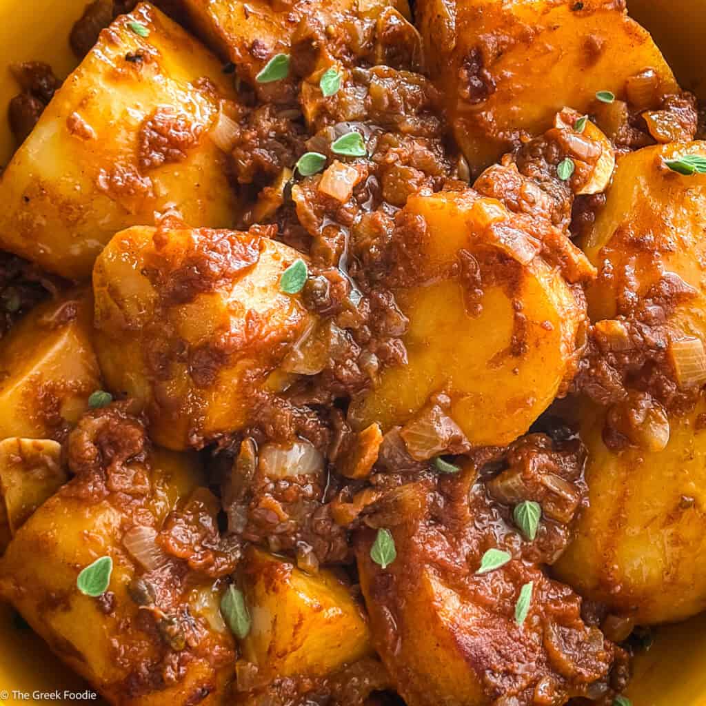 A yellow plate with large potato pieces in a red sauce, a Greek potato stew-patates yahni, with utensils, some herbs on the table and a cloth napkin.