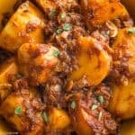 A yellow plate with large potato pieces in a red sauce, a Greek potato stew-patates yahni, with utensils, some herbs on the table and a cloth napkin.