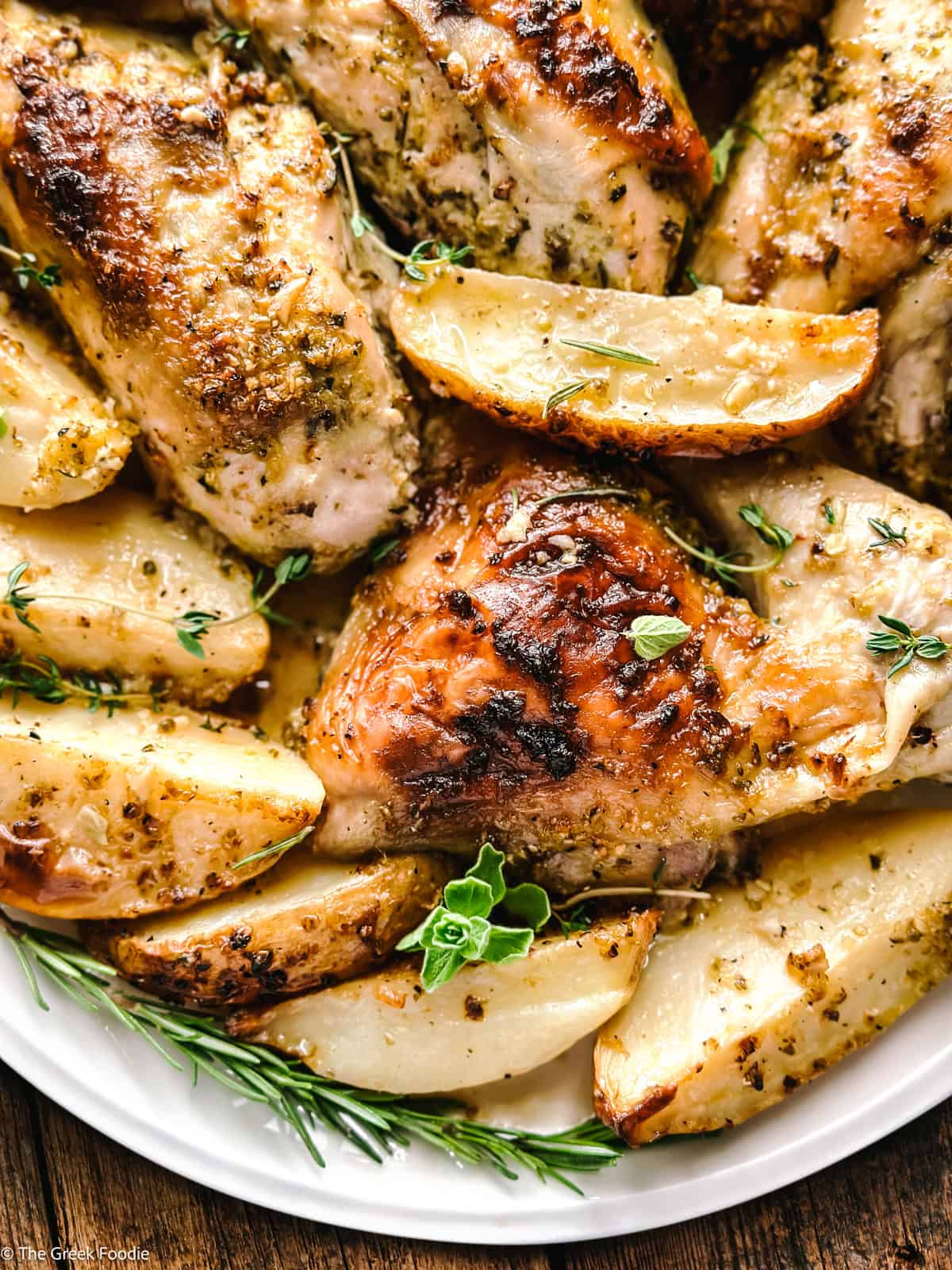 A platter with Greek lemon chicken cut in pieces and roasted potatoes, at the back a citrus squeezer, half a lemon, utensils and a cloth napkin, all on a table.