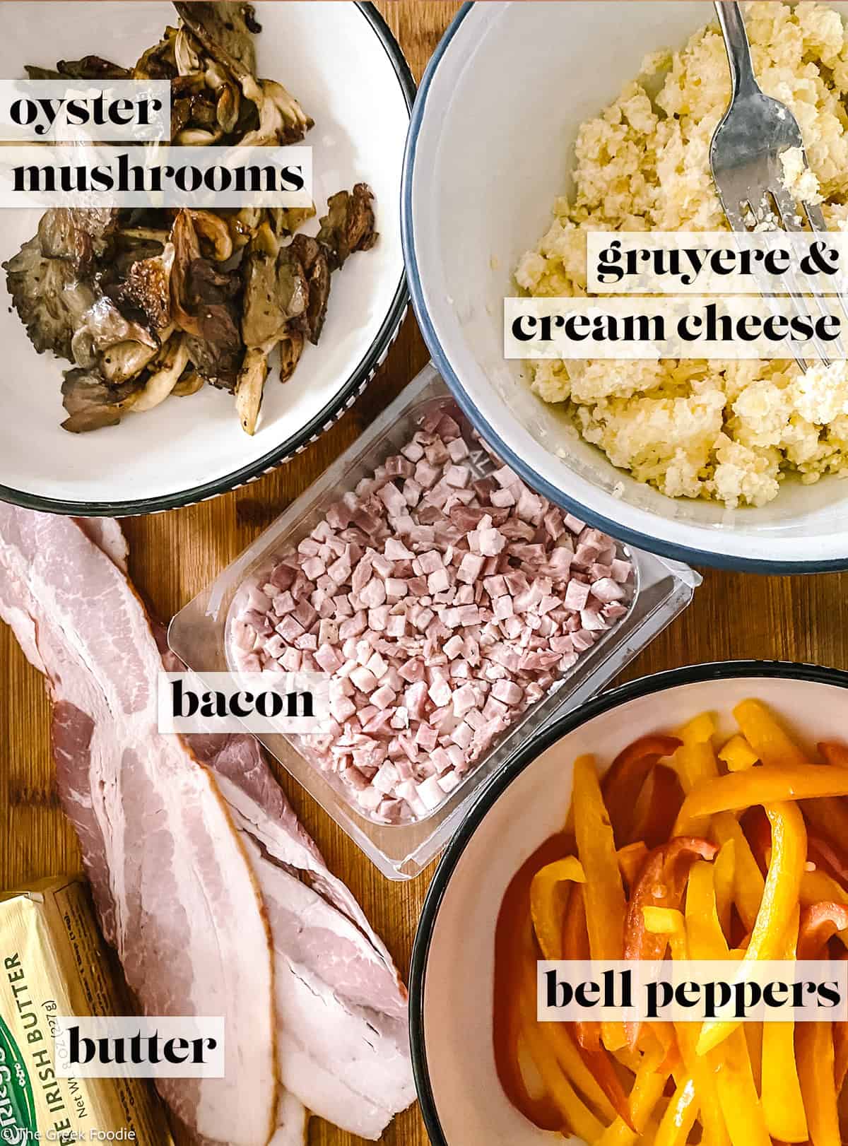 Bowls with mushrooms, cheese, sliced peppers, bacon and chopped bacon and a stick of butter on a wooden surface.