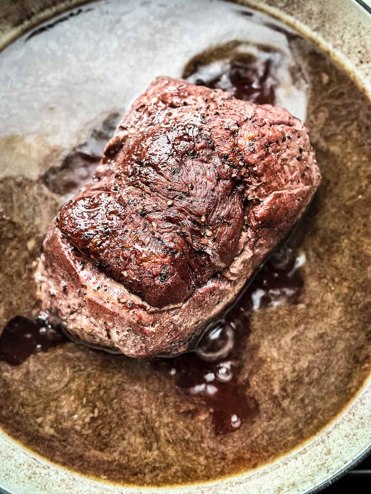 A pork shoulder in a skillet with wine sauce cooking.