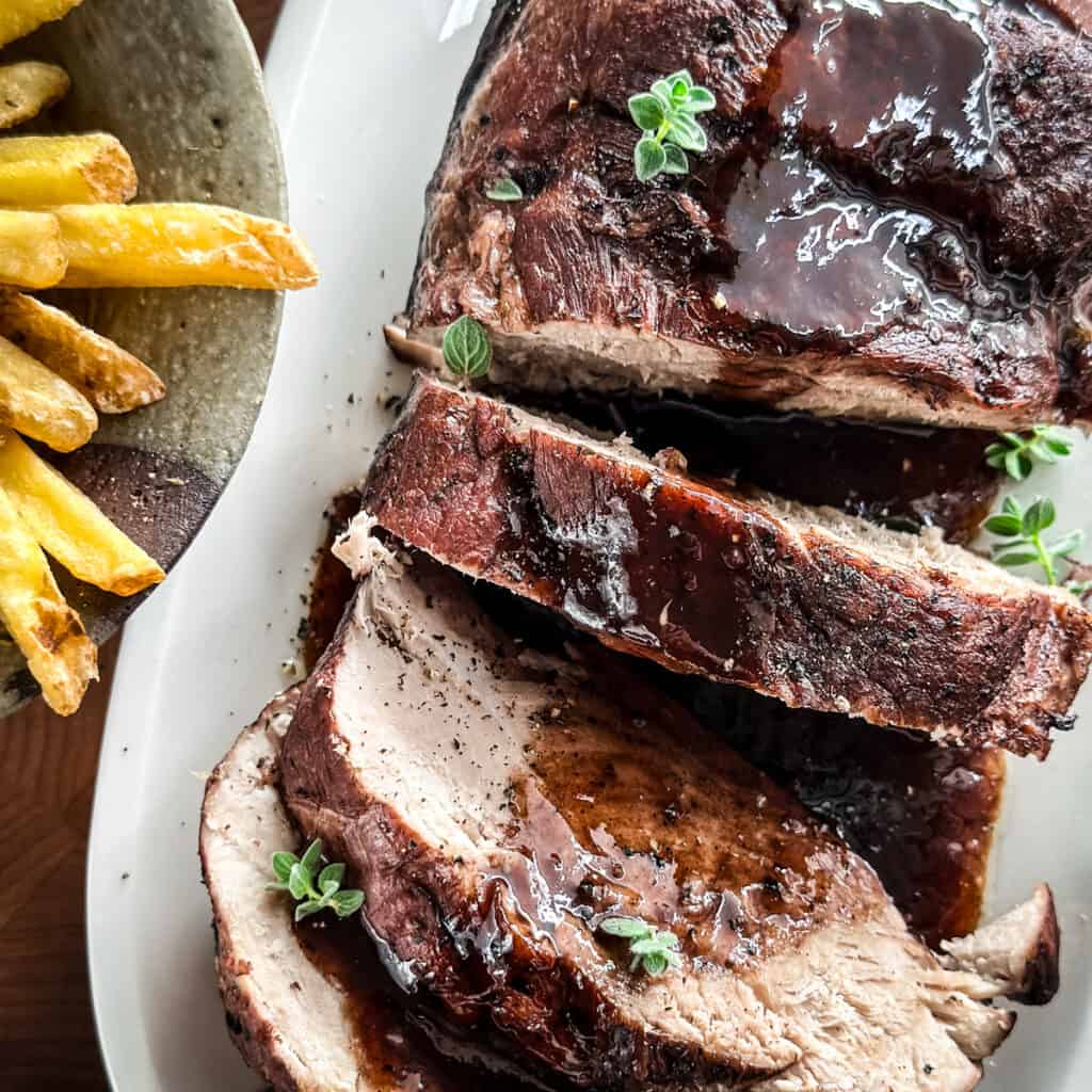 A pork shoulder sliced with wine sauce on a platter next to potato fries.