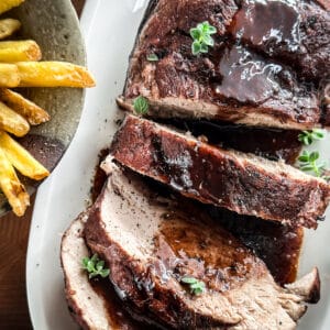 A pork shoulder sliced with wine sauce on a platter next to potato fries.