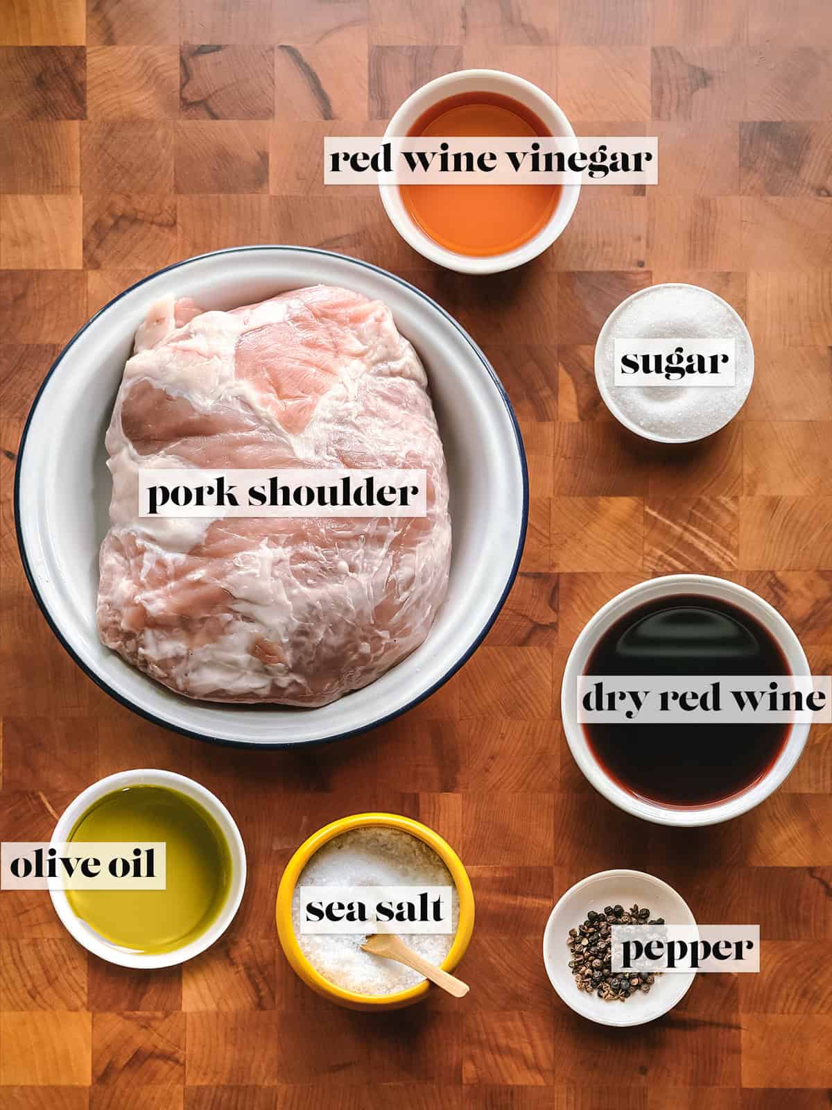 A pork shoulder, sugar, vinegar, wine, olive oil, salt and pepper on a wooden surface, ingredients for red wine pork shoulder braise.