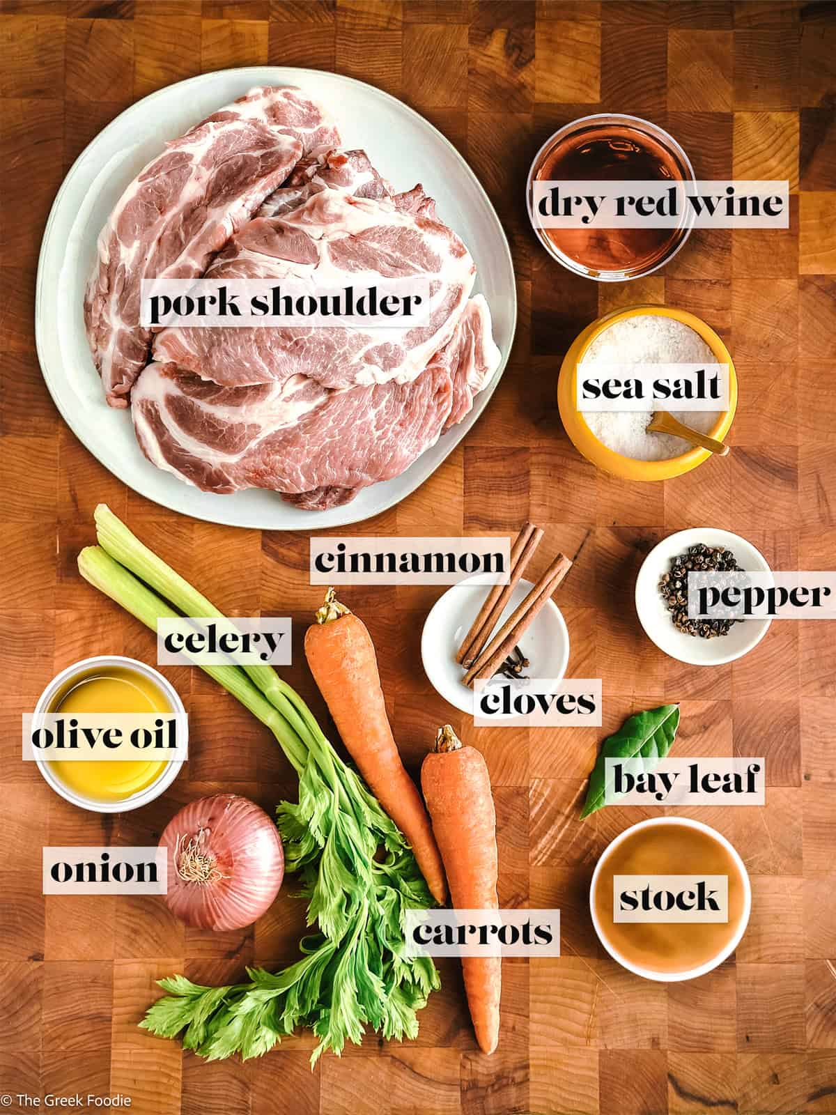 A pork shoulder, wine, olive oil, salt and pepper, carrots and celery and onion, cinnamon and cloves and broth on a wooden surface.