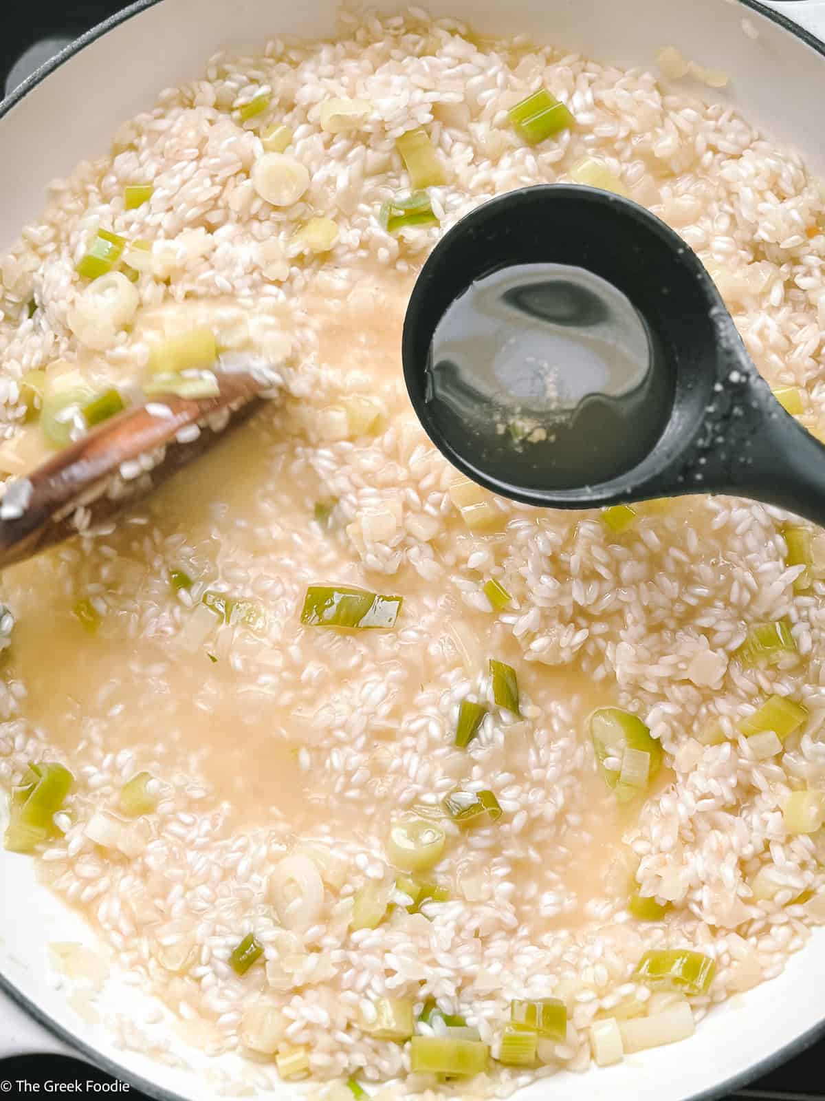 A paddle pouring stock into risotto and a wooden spoon stirring.