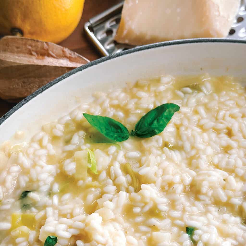 Creamy lemon herb risotto and a spoon in a white large saucepan, a lemon, parmesan at the back.