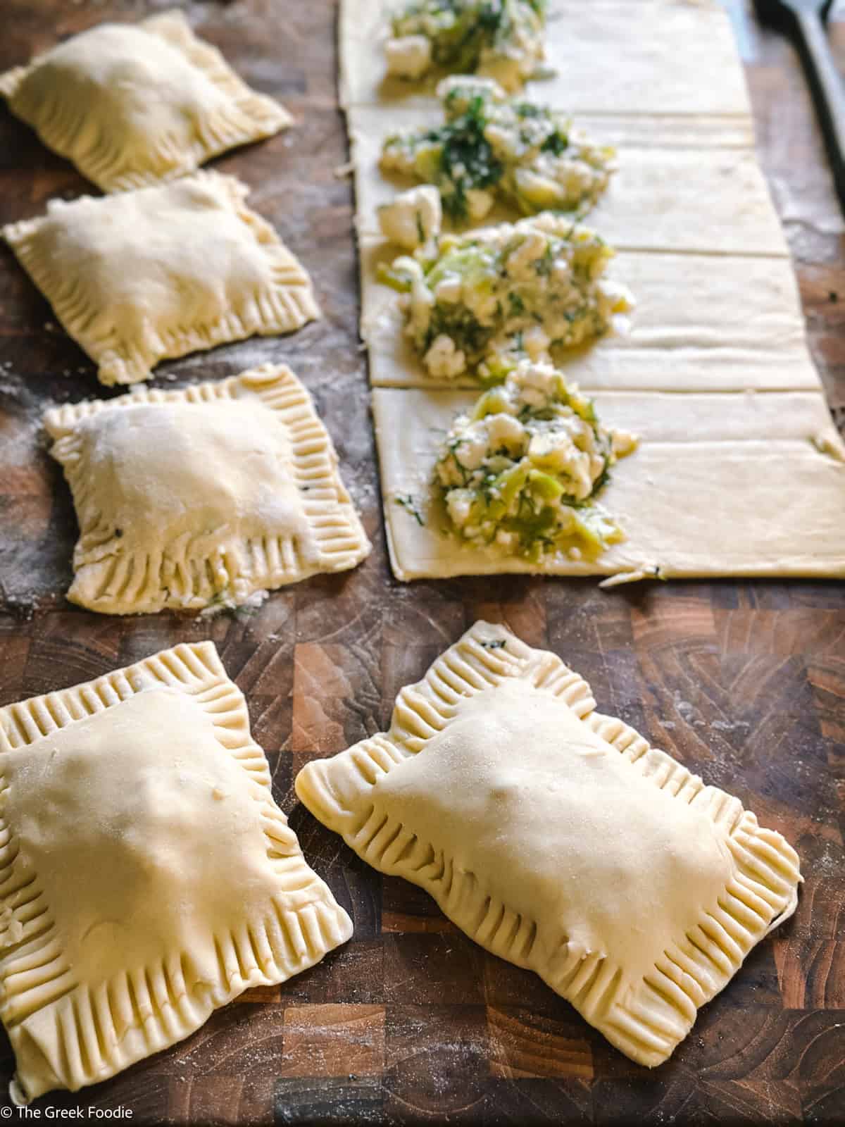Puff pastry strips with feta cheese filling on them, and a few sealed puff pastries on the front, on a wooden board.