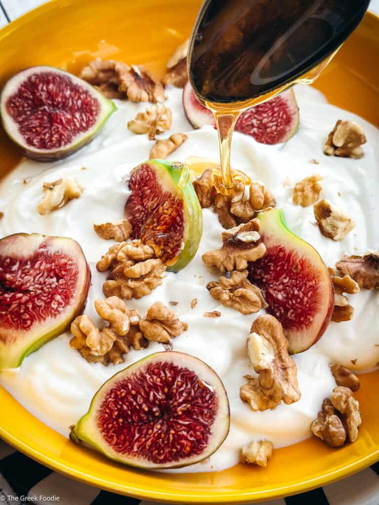 A spoon drizzling honey on a plate with yogurt, sliced figs, and walnuts.