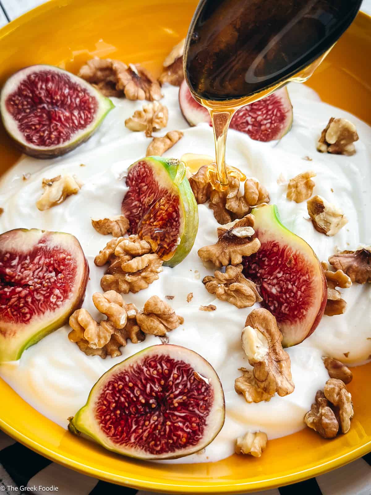 A spoon drizzling honey on a plate with yogurt, sliced figs, and walnuts.