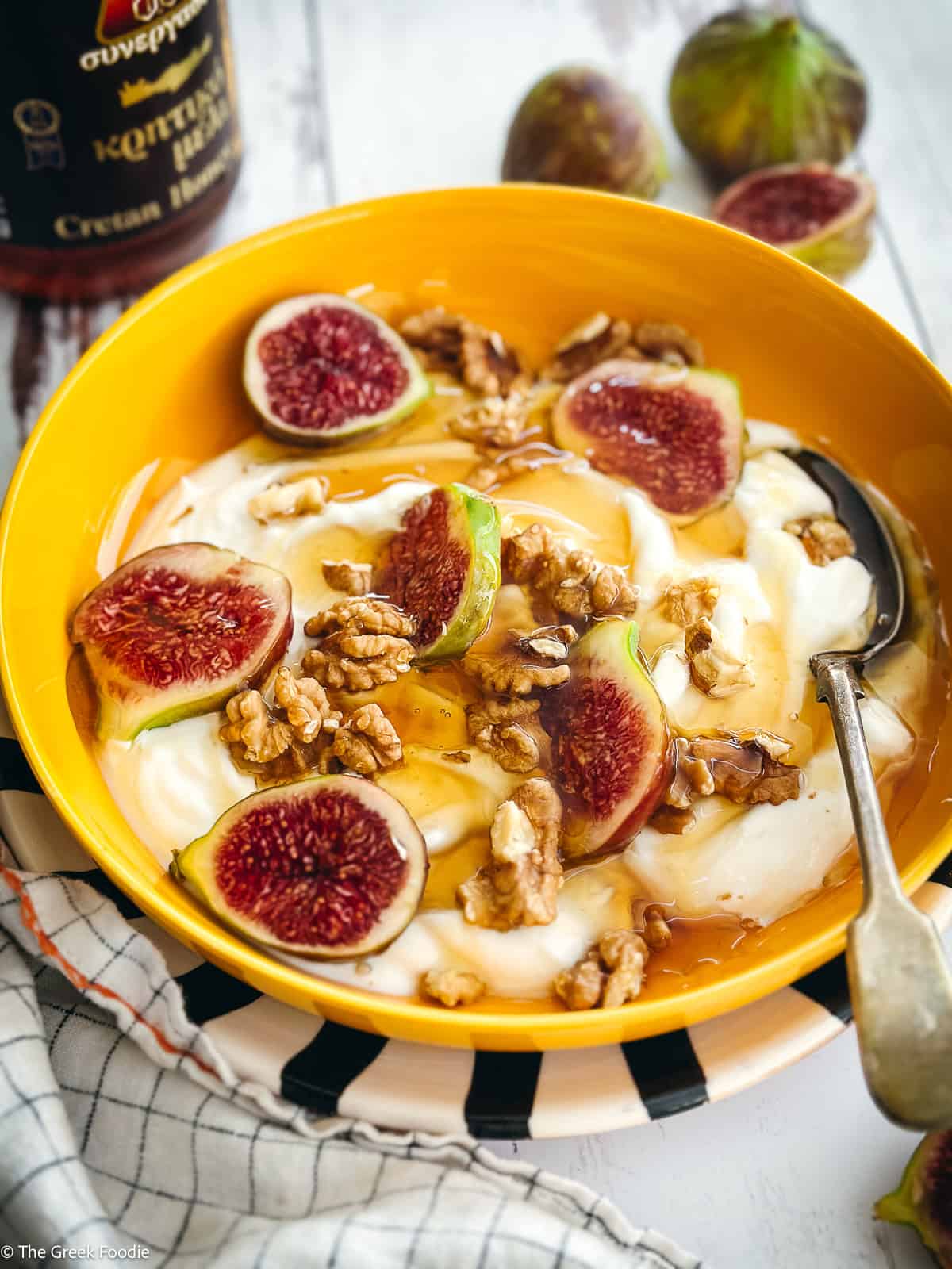 A plate with yogurt, sliced figs, walnuts and honey and a spoon. At the back figs and a jar with honey.
