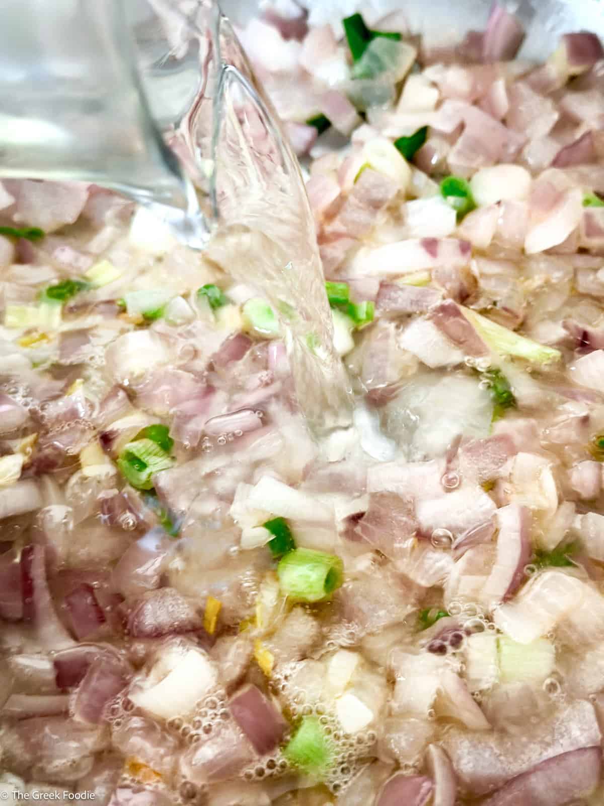 A glass cup pouring white wine in to a pan with diced onions.