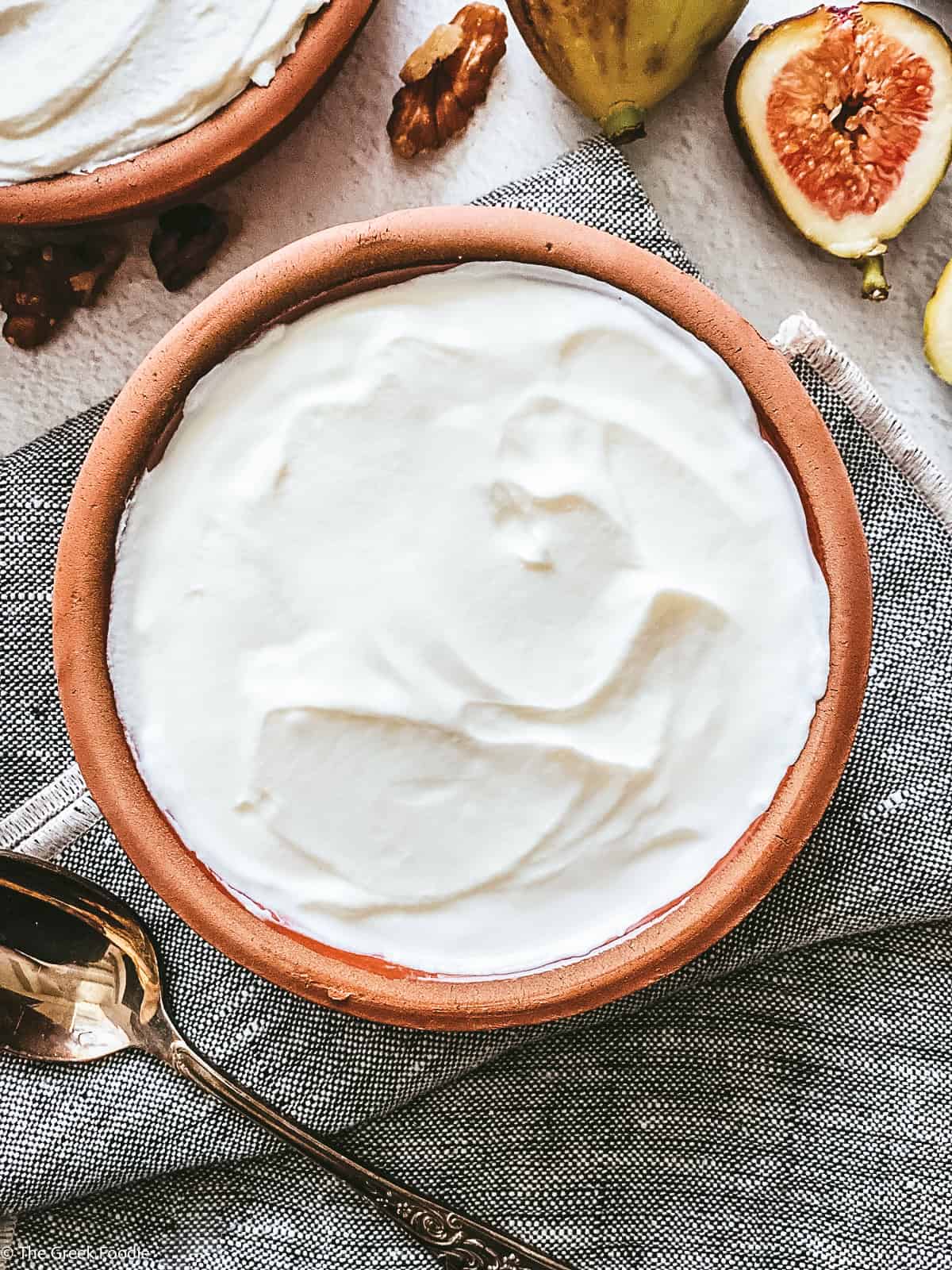 A ceramic bowl with yogurt and a spoon on a cloth napkin, fig halves above it and a detail of another bowl.