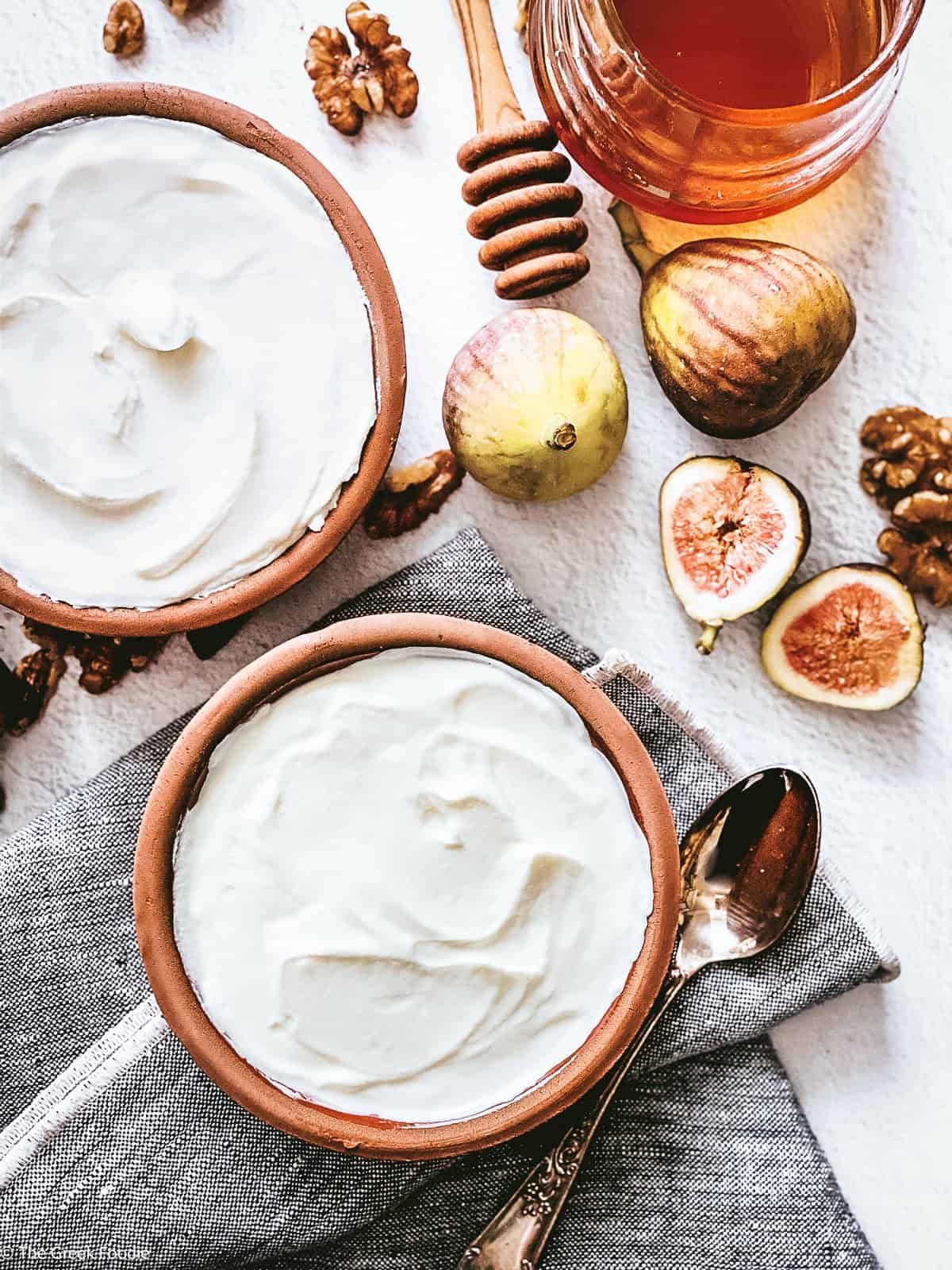 Two ceramic bowls with yogurt, a spoon on a cloth napkin, fig halves, walnuts, a honey dipper and a jar with honey. and a detail of another bowl.