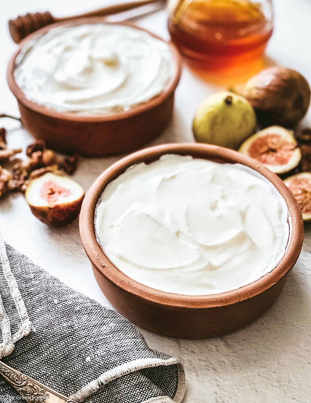 Two ceramic bowls with homemade greek yogurt, a spoon on a cloth napkin, fig halves, walnuts, a honey dipper and a jar with honey. and a detail of another bowl.