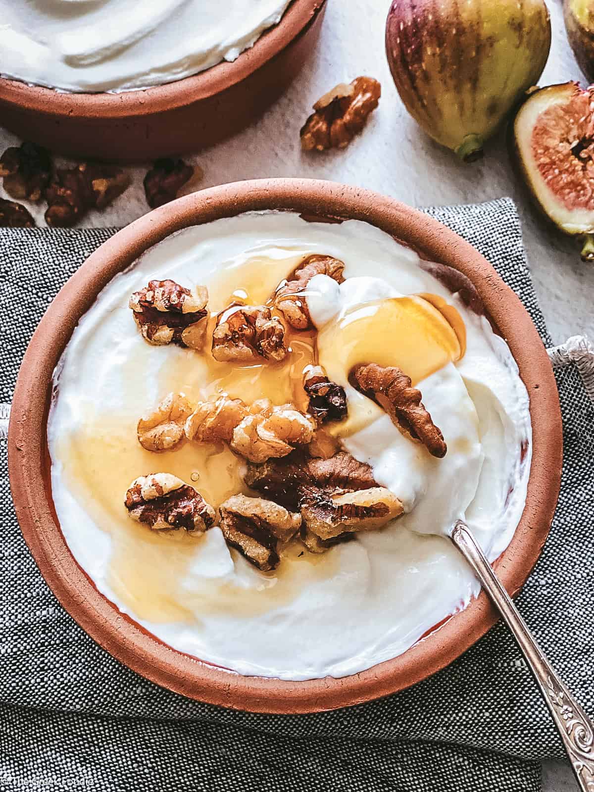 A bowl with yogurt, a honey dipper dripping honey, a spoon on a cloth napkin, fig halves, walnuts, and a detail of another bowl.