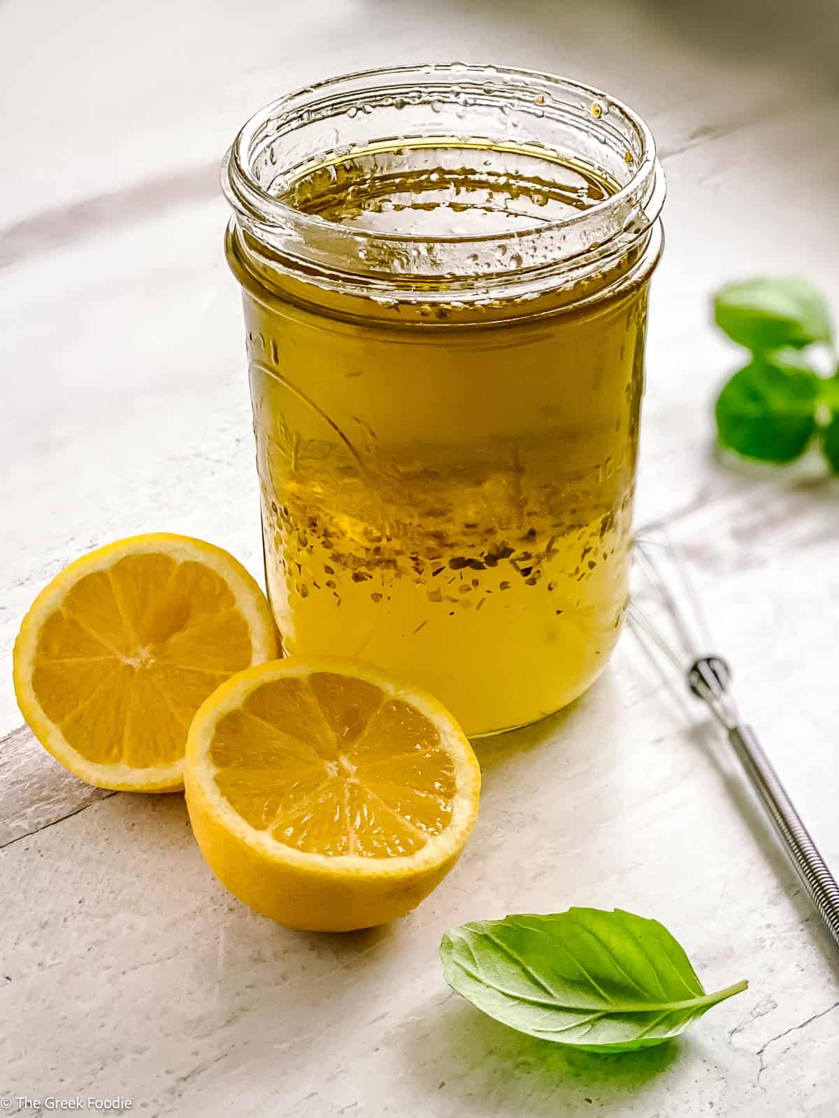A glass jar with lemon and olive oil dressing-ladolemono, two lemon halves, a whisk and some fresh basil.