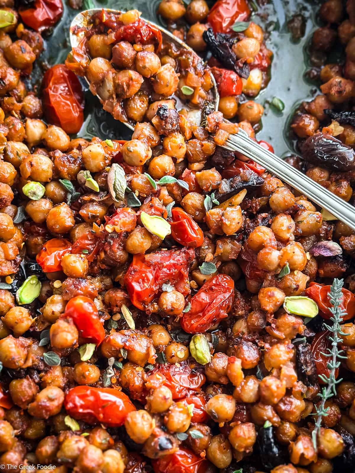 Warm chickpea salad with tomatoes and pistachios in a blue baking pan with a spoon.