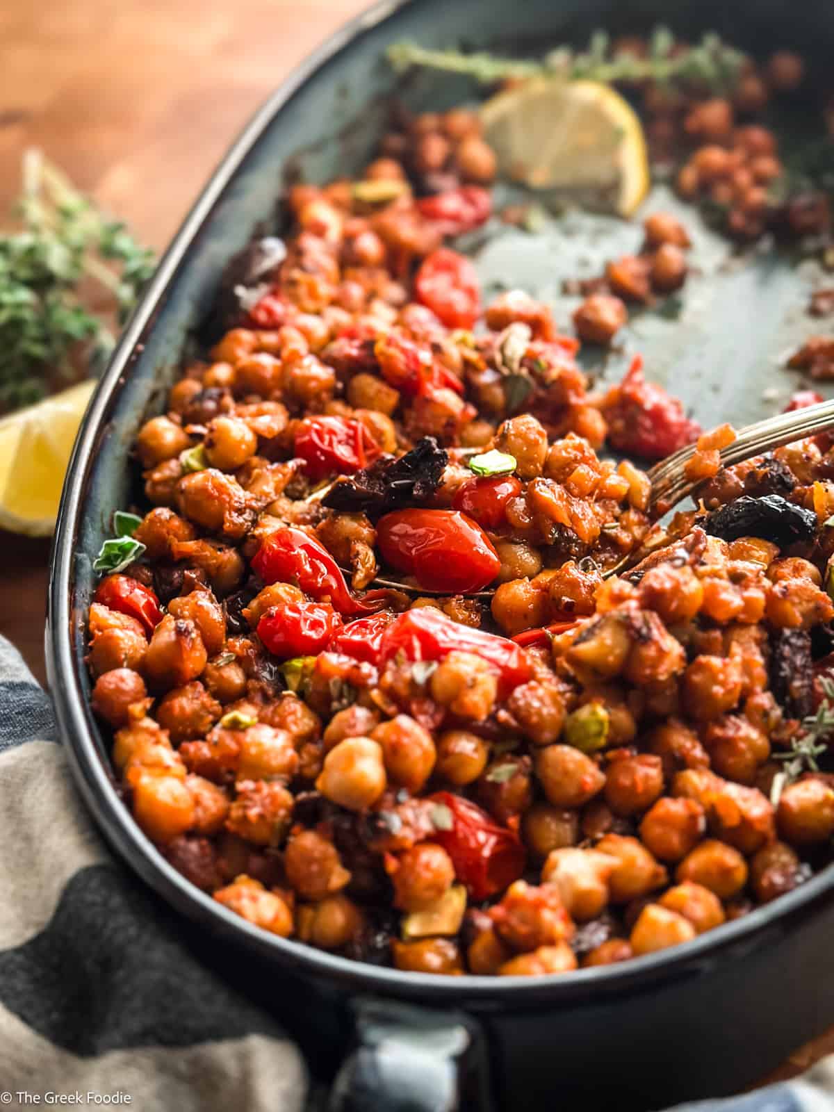 Warm chickpea salad with tomatoes and pistachios in a blue baking pan with a spoon, on a table.