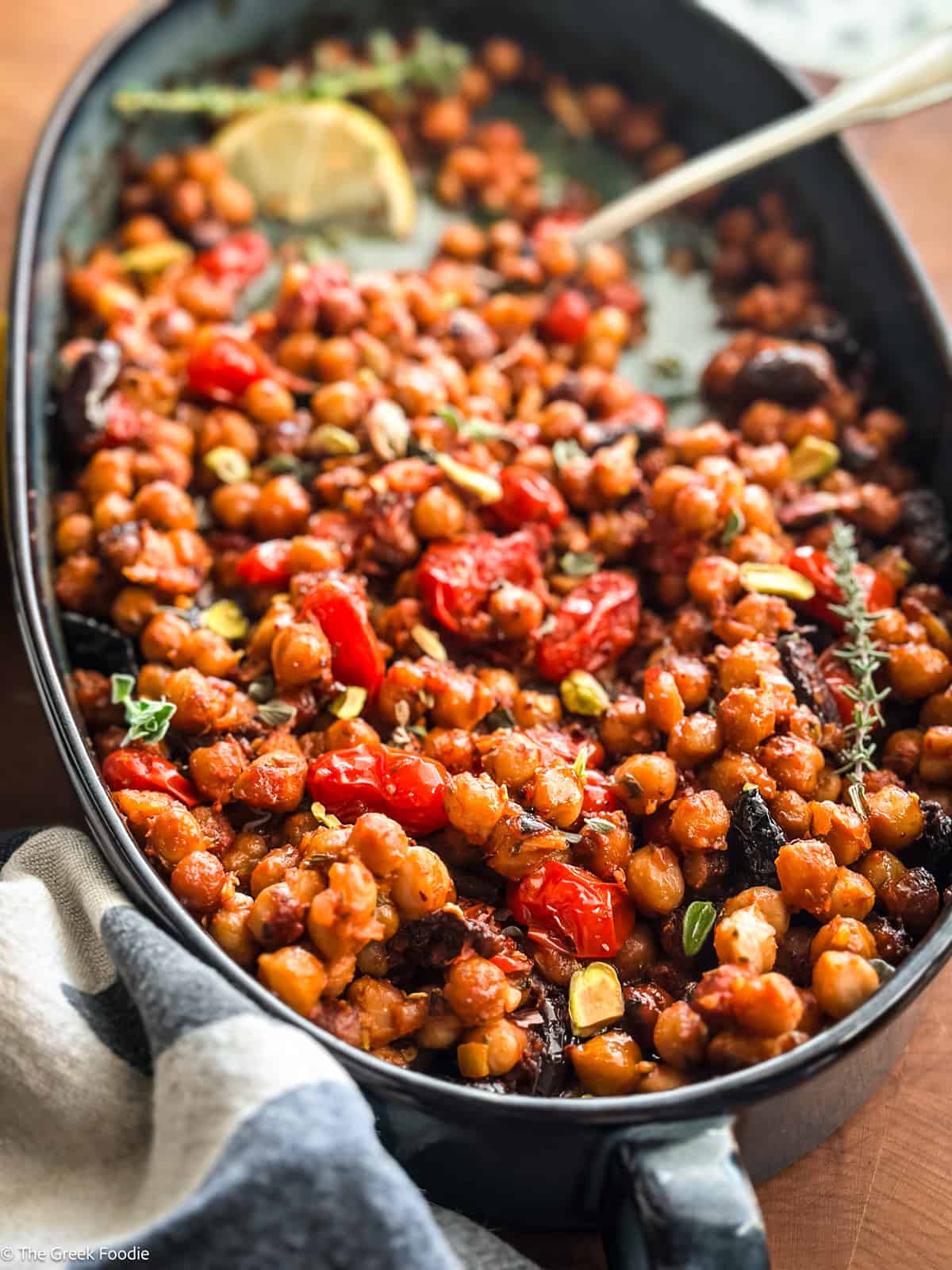 Warm chickpea salad with tomatoes and pistachios in a blue baking pan with a spoon, on a table.