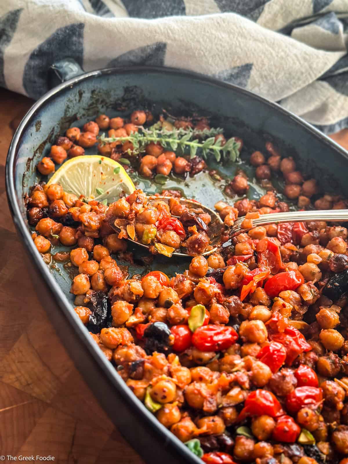 Warm chickpea salad with tomatoes and pistachios in a blue baking pan with a spoon, on a table.