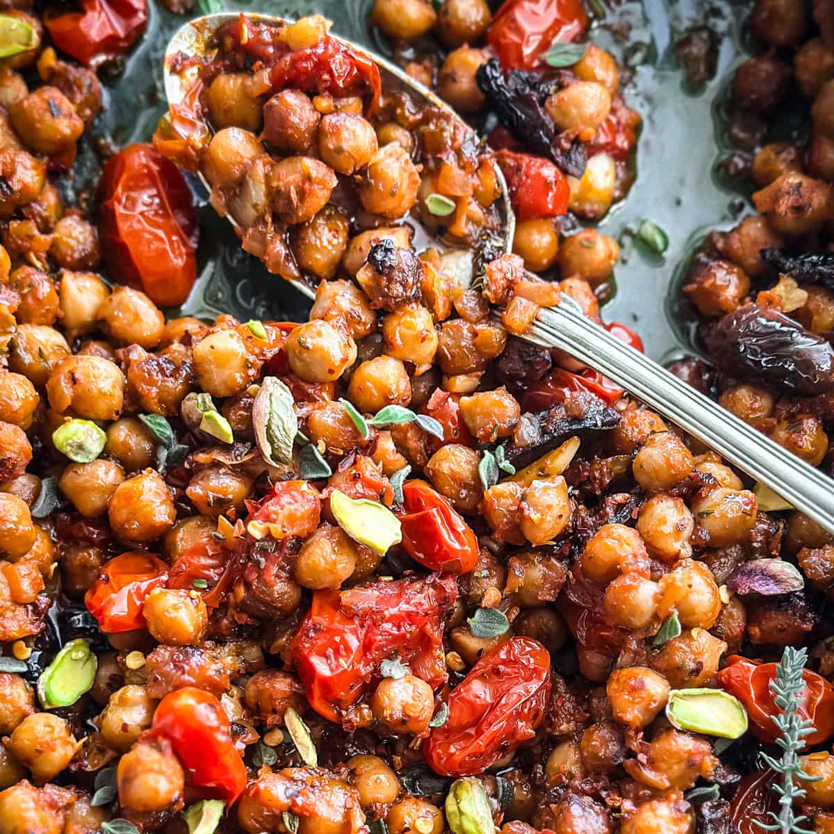 Warm chickpea salad with tomatoes and pistachios in a blue baking pan with a spoon.