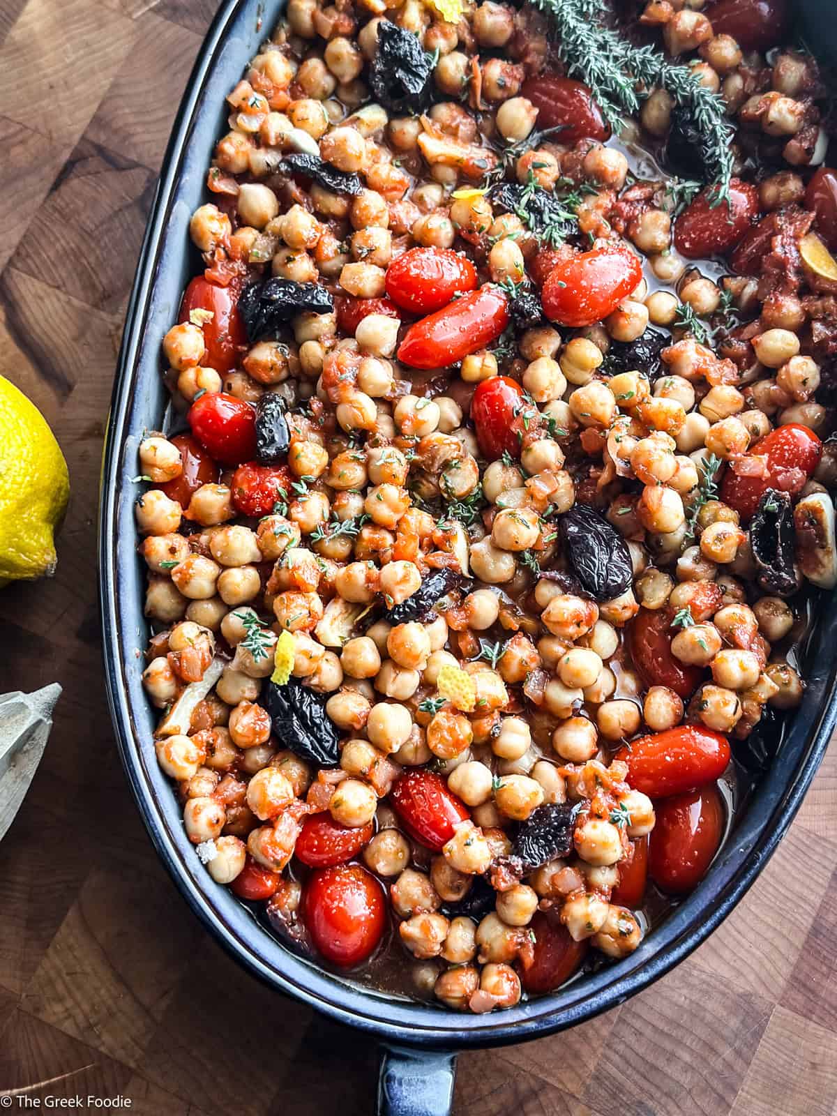 Prepped chickpea salad with tomatoes and pistachios in a blue baking pan with a spoon, on a table.