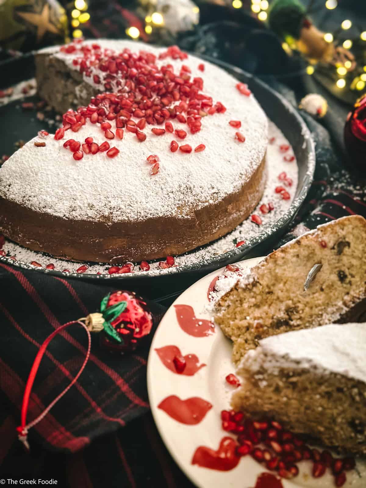 A vasilopita cake at the back, two cake pieces on a plate at the front.