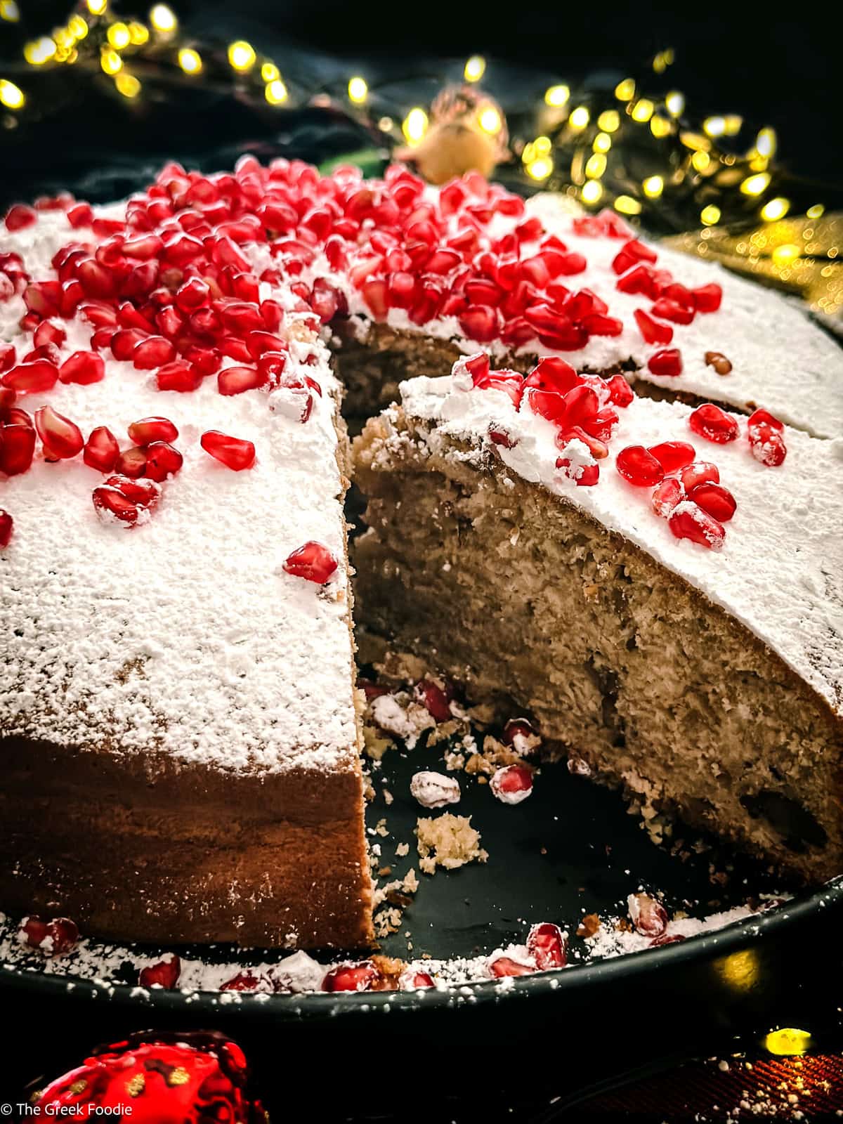 A Vasilopita cake-Greek new years cake decorated with pomegranate seeds with a piece cut off on a platter, behind and around it Christmas lights and decorations.