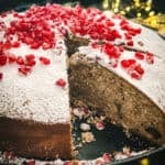 A Vasilopita cake-Greek new years cake decorated with pomegranate seeds with a piece cut off on a platter, behind and around it Christmas lights and decorations.