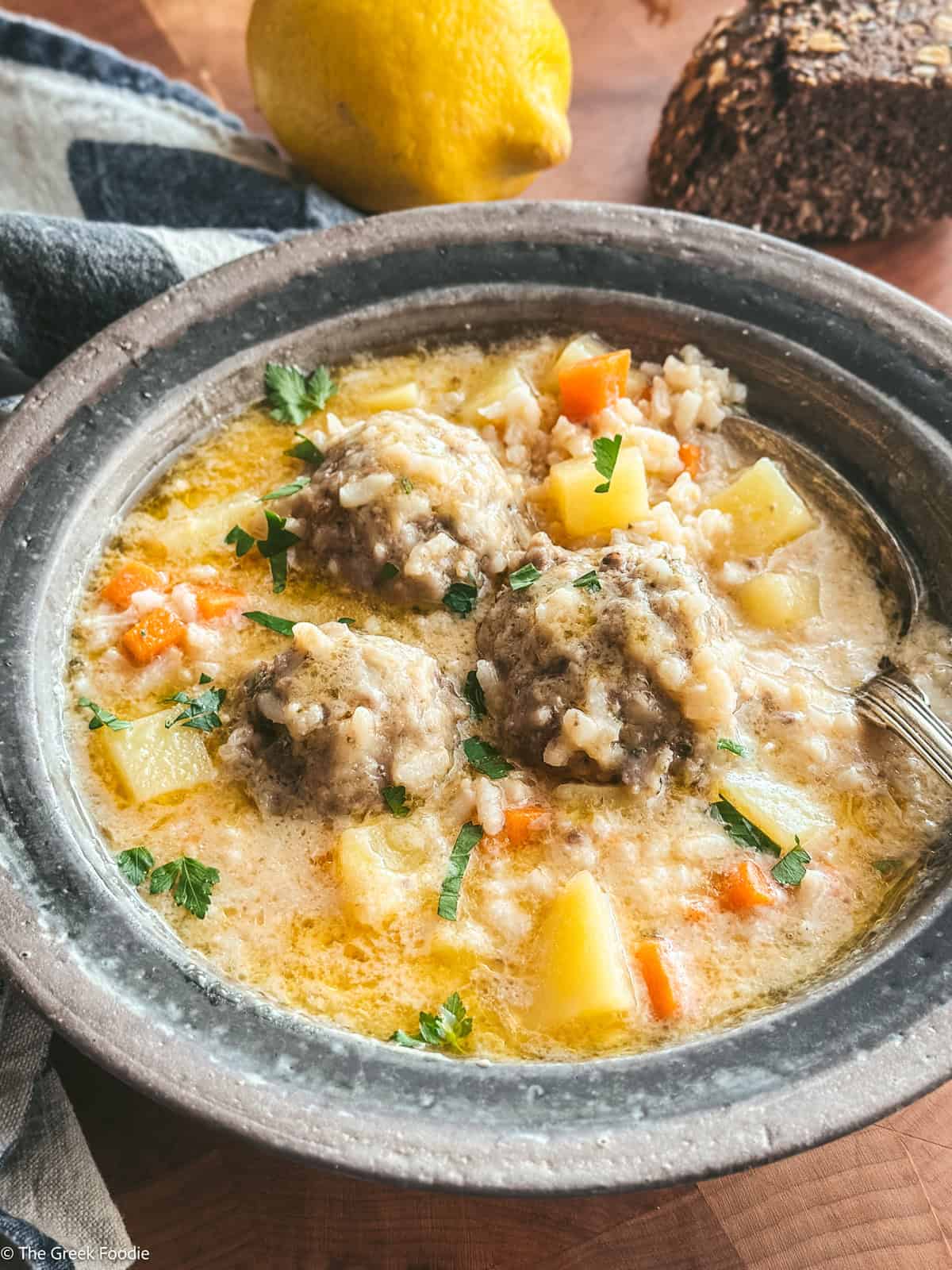 Youvarlakia meatballs with rice, broth in a plate with a spoon, on a table with bread and a lemon.