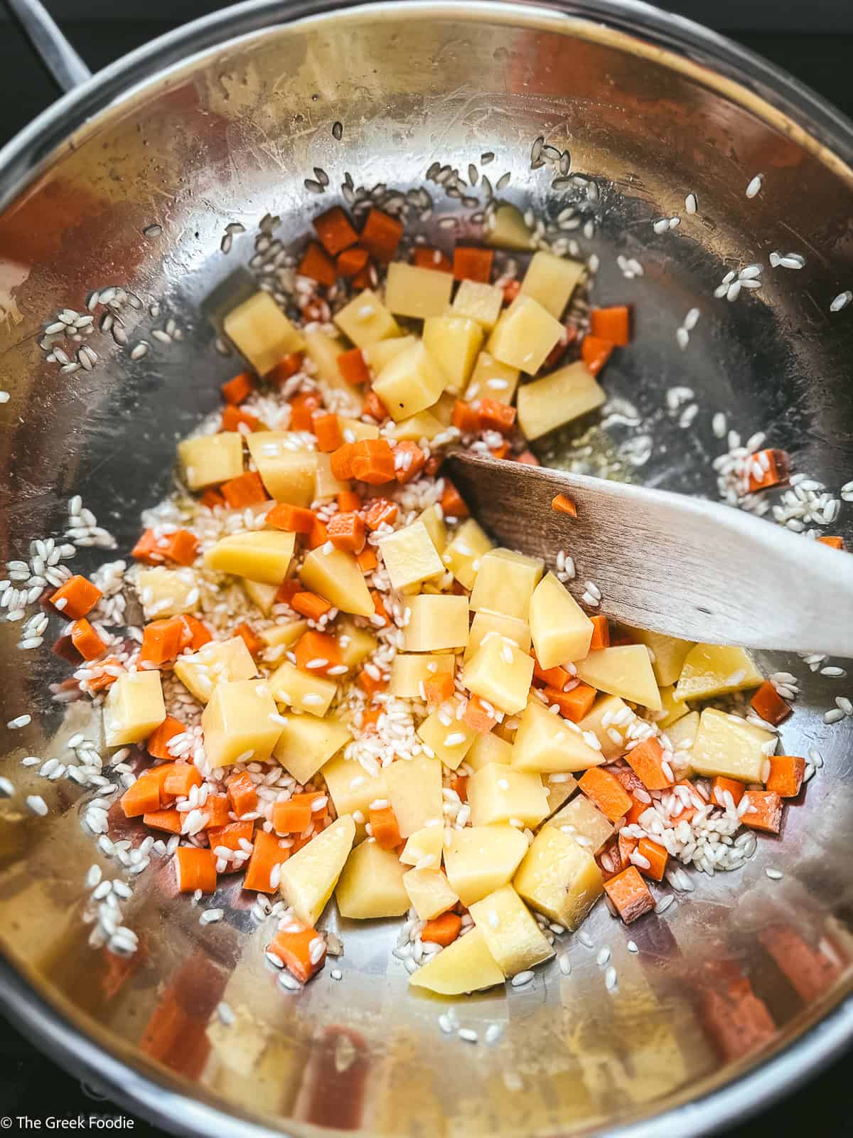 Potato cubes and carrot cubes with rice cooking in a pan with a wooden spoon.
