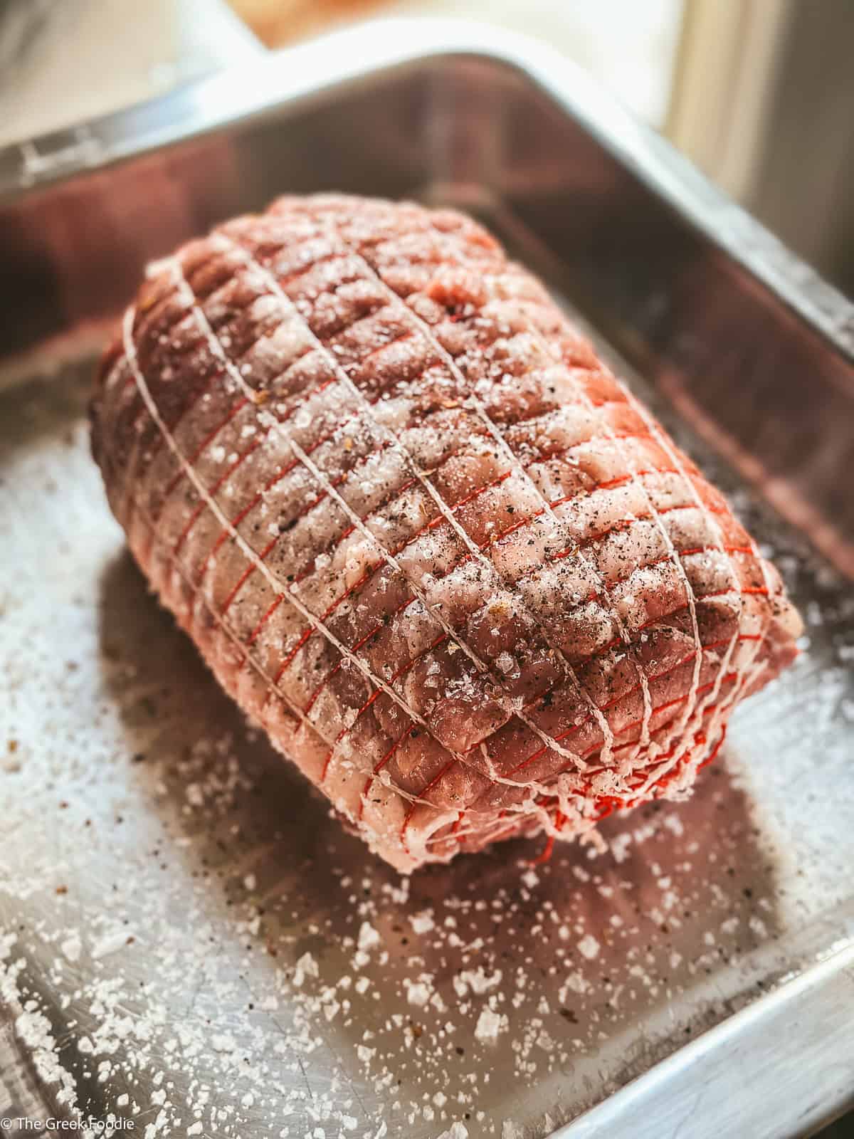 A pork shoulder wrapped in a net, seasoned with sea salt and pepper in a baking tray.